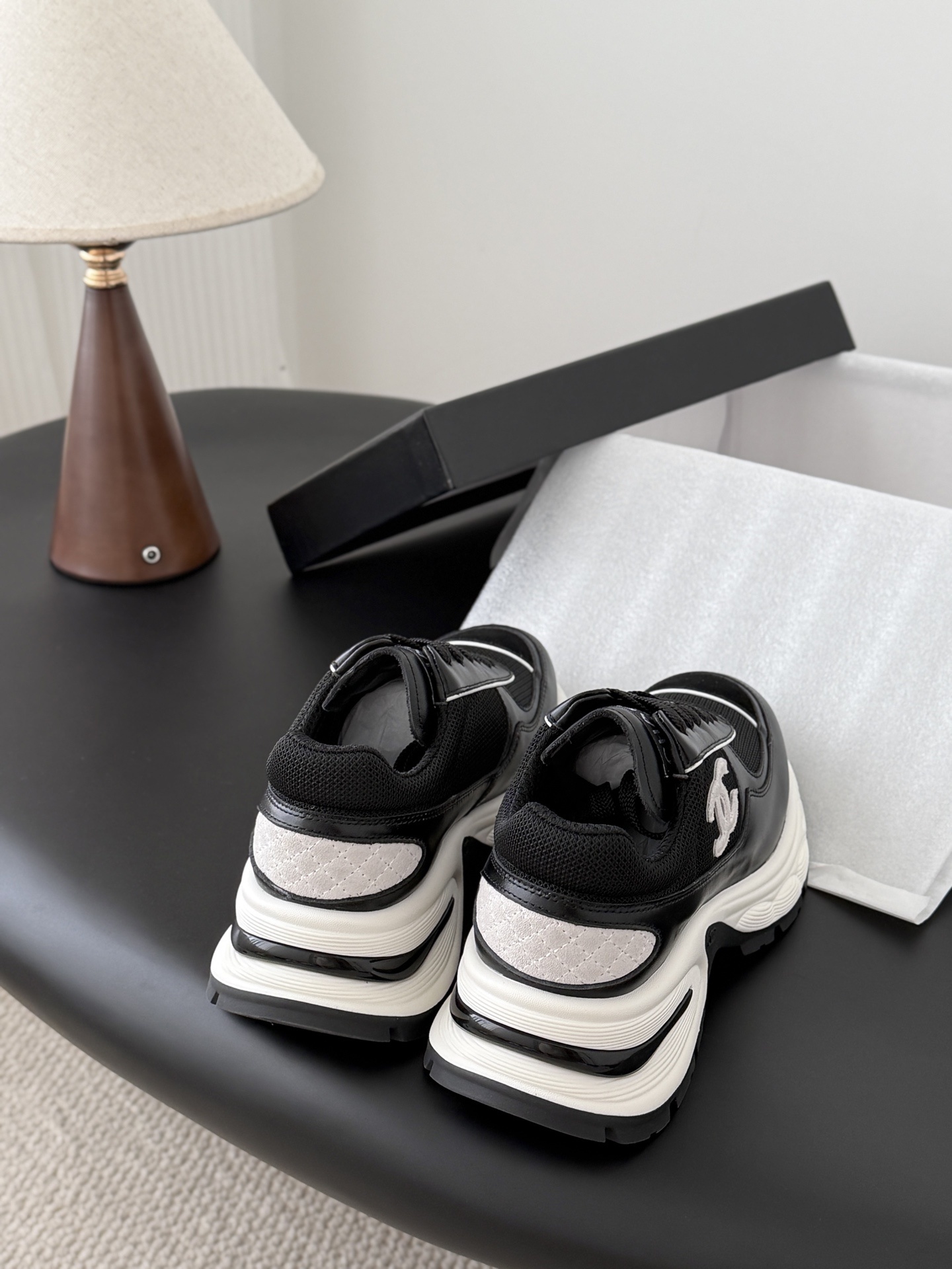 A pair of black and white Chanel chunky leather sneakers featuring the signature CC logo on a black surface next to a branded box. - Hình 3