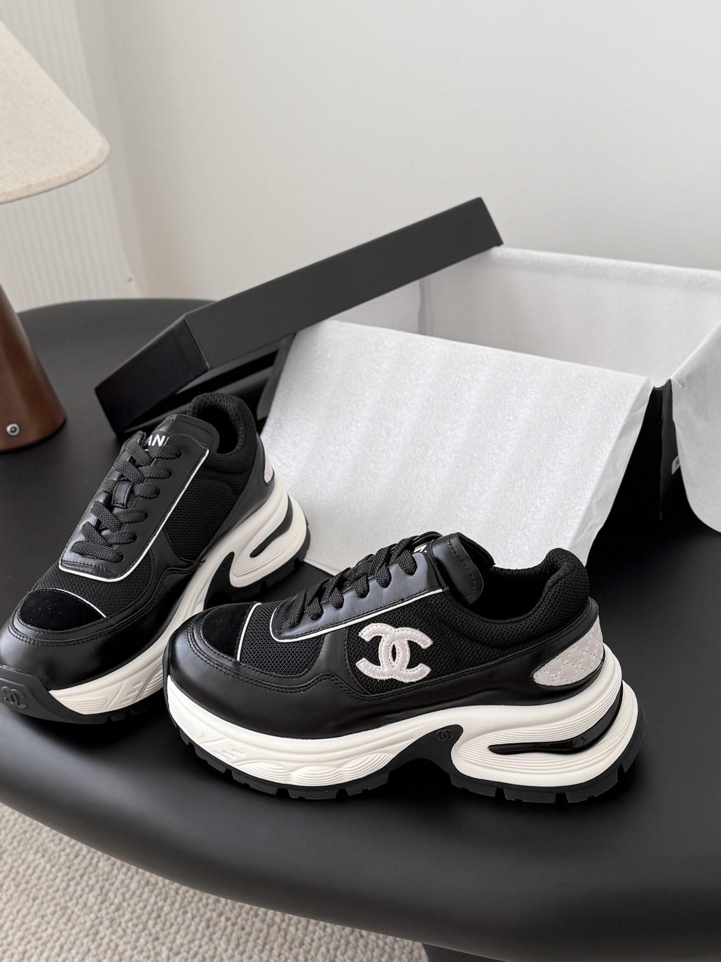 A pair of black and white Chanel chunky leather sneakers featuring the signature CC logo on a black surface next to a branded box. - Hình 2