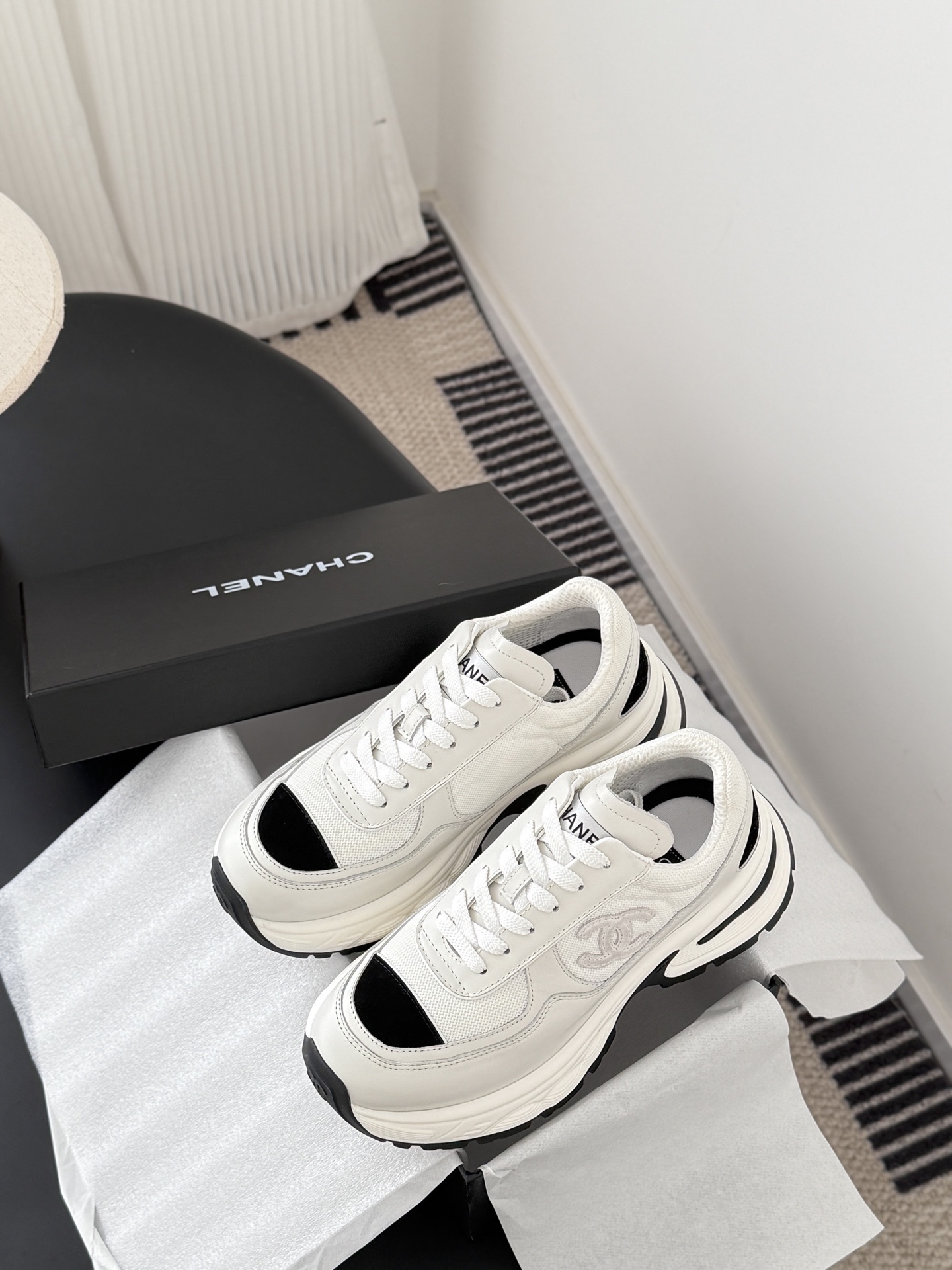 A pair of Chanel white chunky sneakers with black accents and interlocking CC logo displayed on a black surface next to a Chanel shoe box. - Hình 9