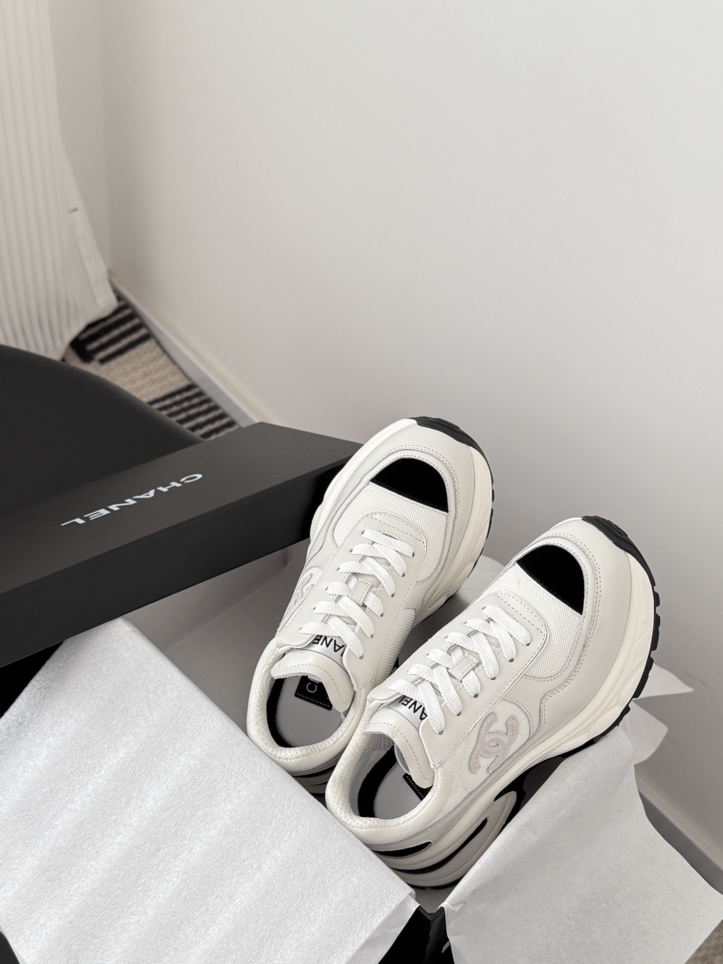 A pair of Chanel white chunky sneakers with black accents and interlocking CC logo displayed on a black surface next to a Chanel shoe box. - Hình 8
