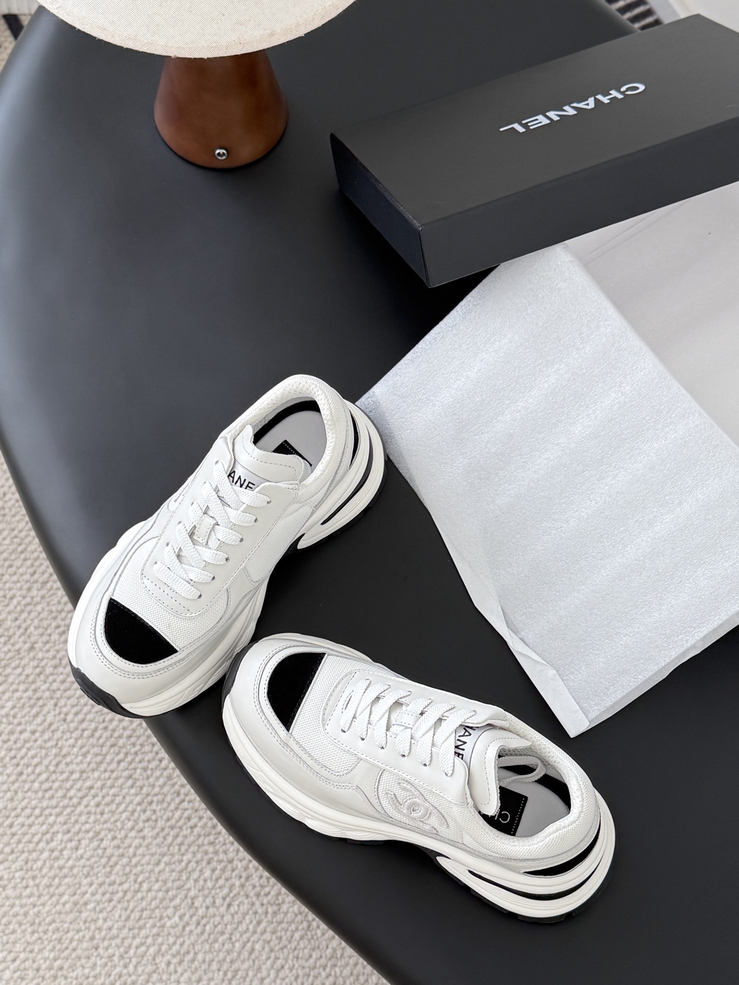 A pair of Chanel white chunky sneakers with black accents and interlocking CC logo displayed on a black surface next to a Chanel shoe box. - Hình 6