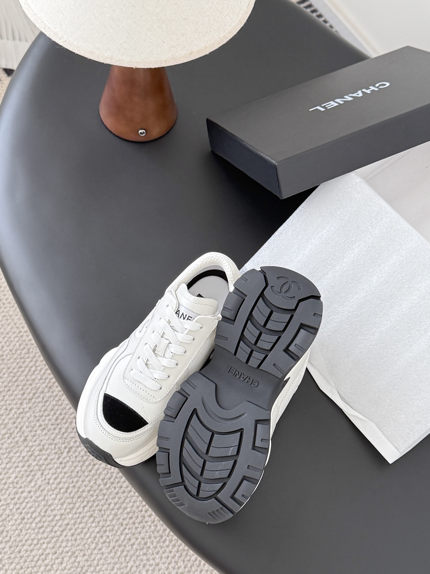 A pair of Chanel white chunky sneakers with black accents and interlocking CC logo displayed on a black surface next to a Chanel shoe box. - Hình 5