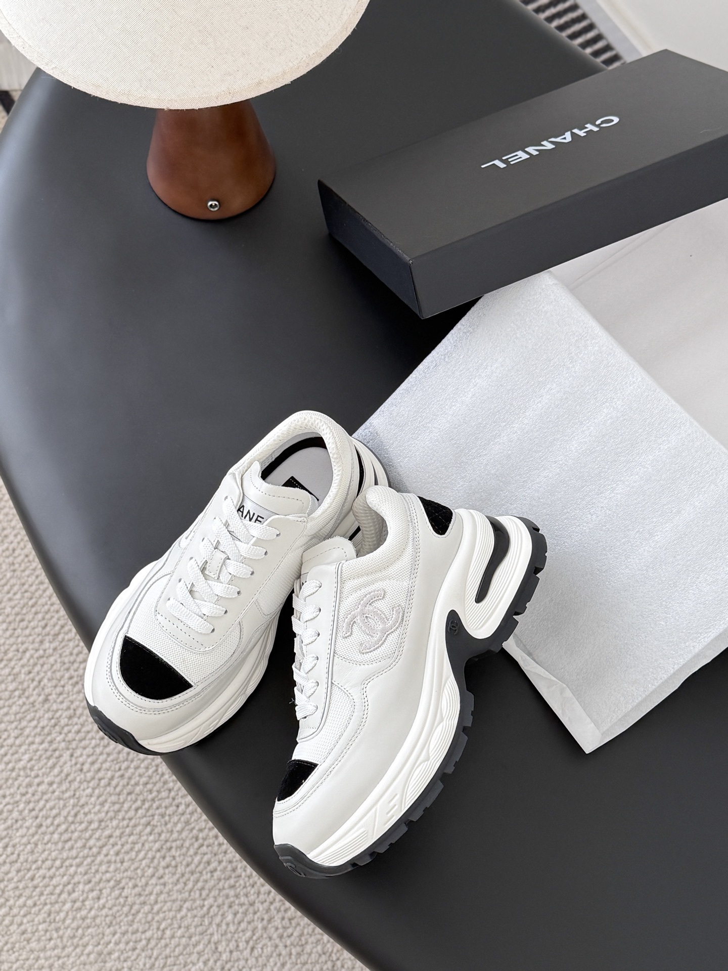 A pair of Chanel white chunky sneakers with black accents and interlocking CC logo displayed on a black surface next to a Chanel shoe box. - Hình 4