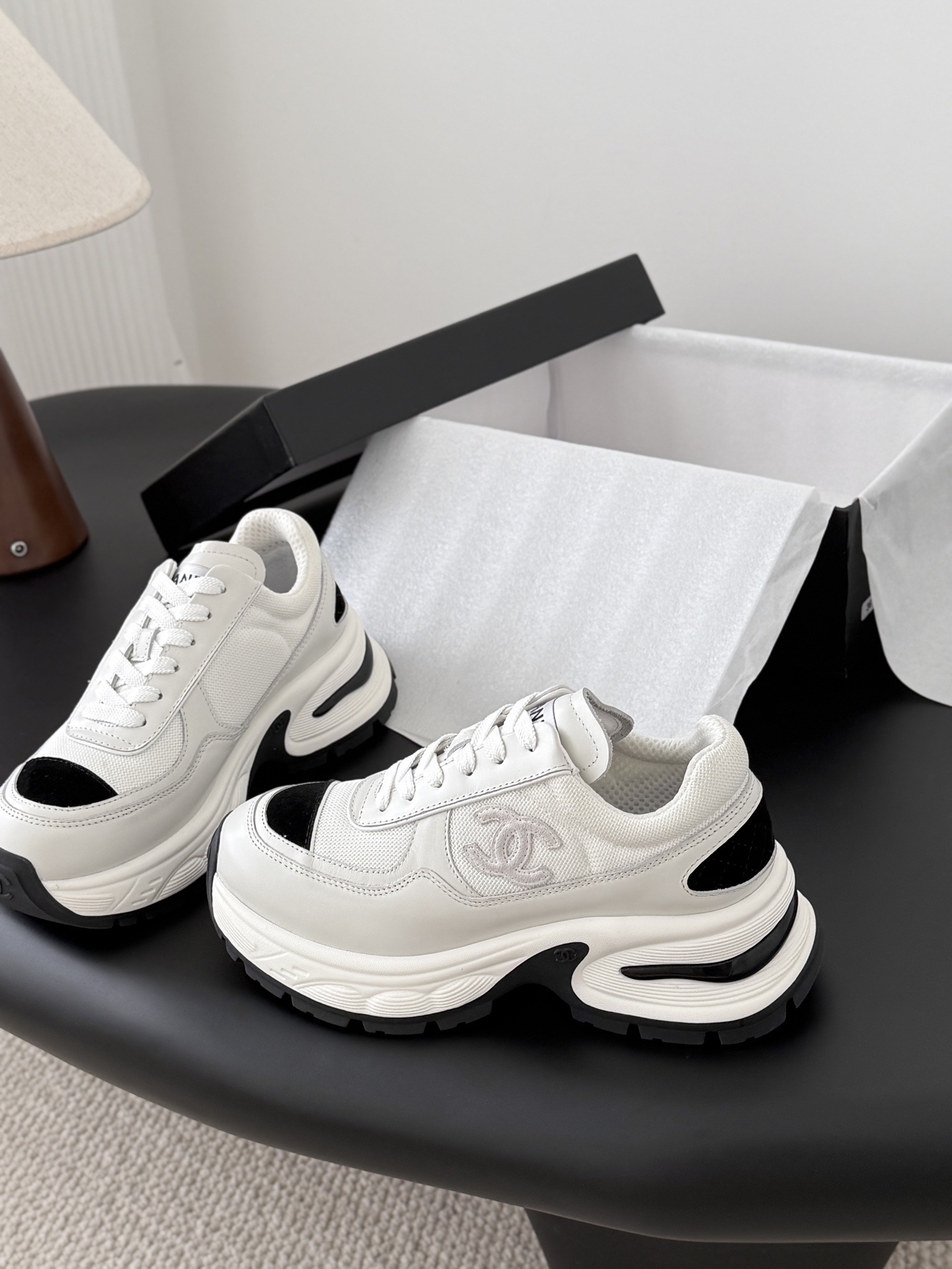 A pair of Chanel white chunky sneakers with black accents and interlocking CC logo displayed on a black surface next to a Chanel shoe box. - Hình 2
