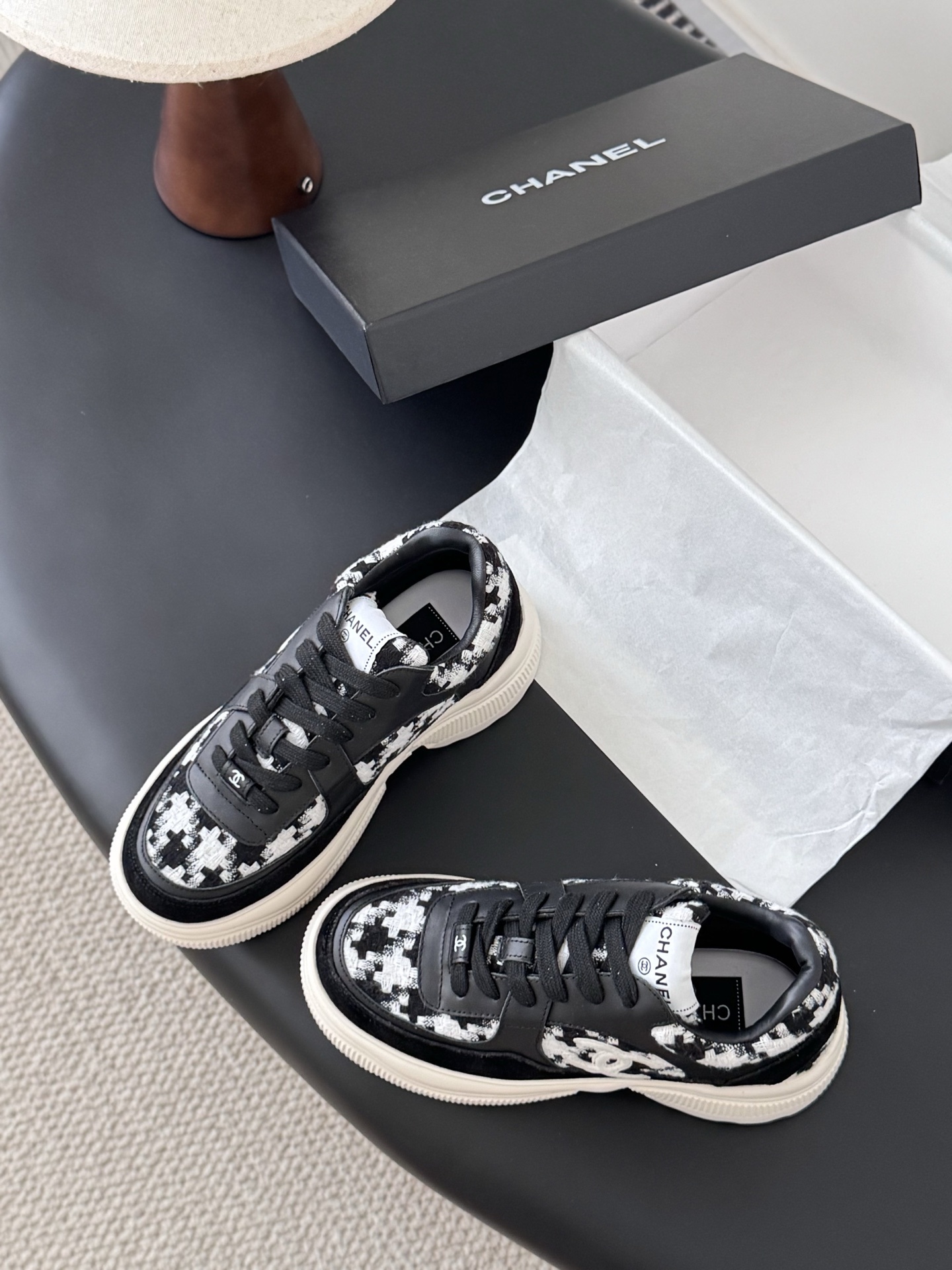 A pair of Chanel black and white houndstooth tweed sneakers with chunky white soles displayed next to a Chanel shoe box. - Hình 6