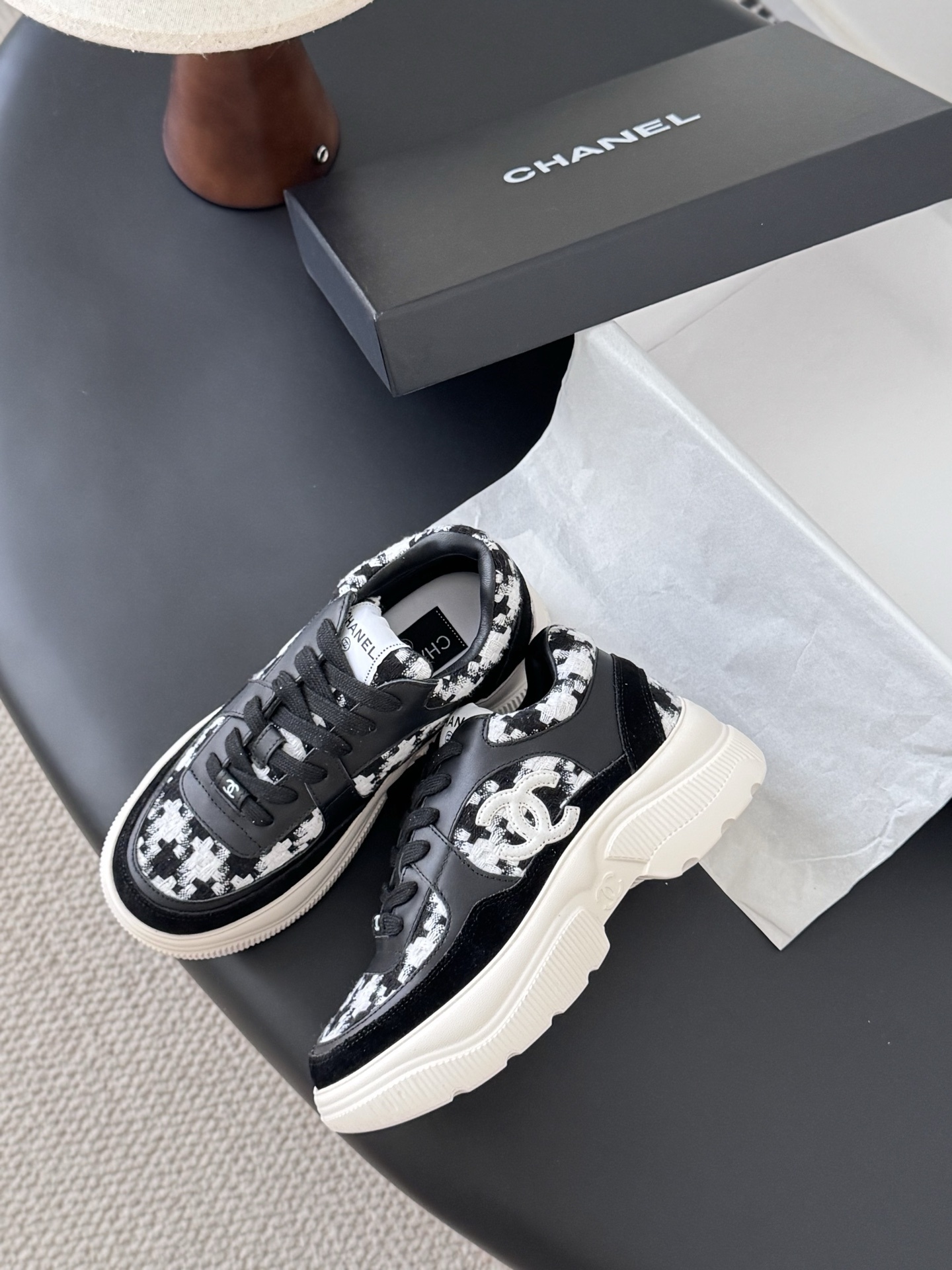A pair of Chanel black and white houndstooth tweed sneakers with chunky white soles displayed next to a Chanel shoe box. - Hình 4