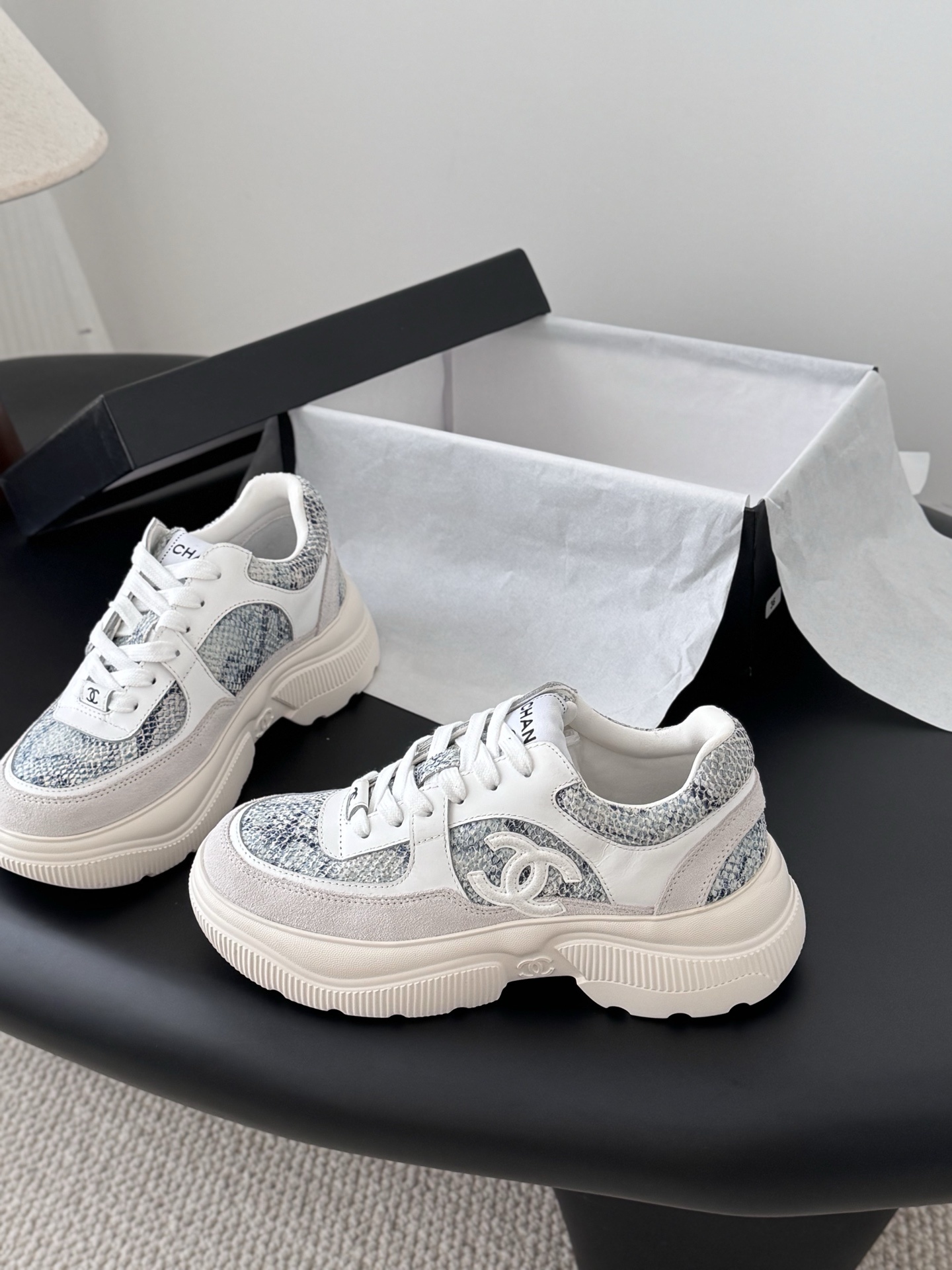 A pair of Chanel white chunky sneakers with blue snakeskin textured panels and the CC logo, displayed next to a black Chanel box. - Hình 2
