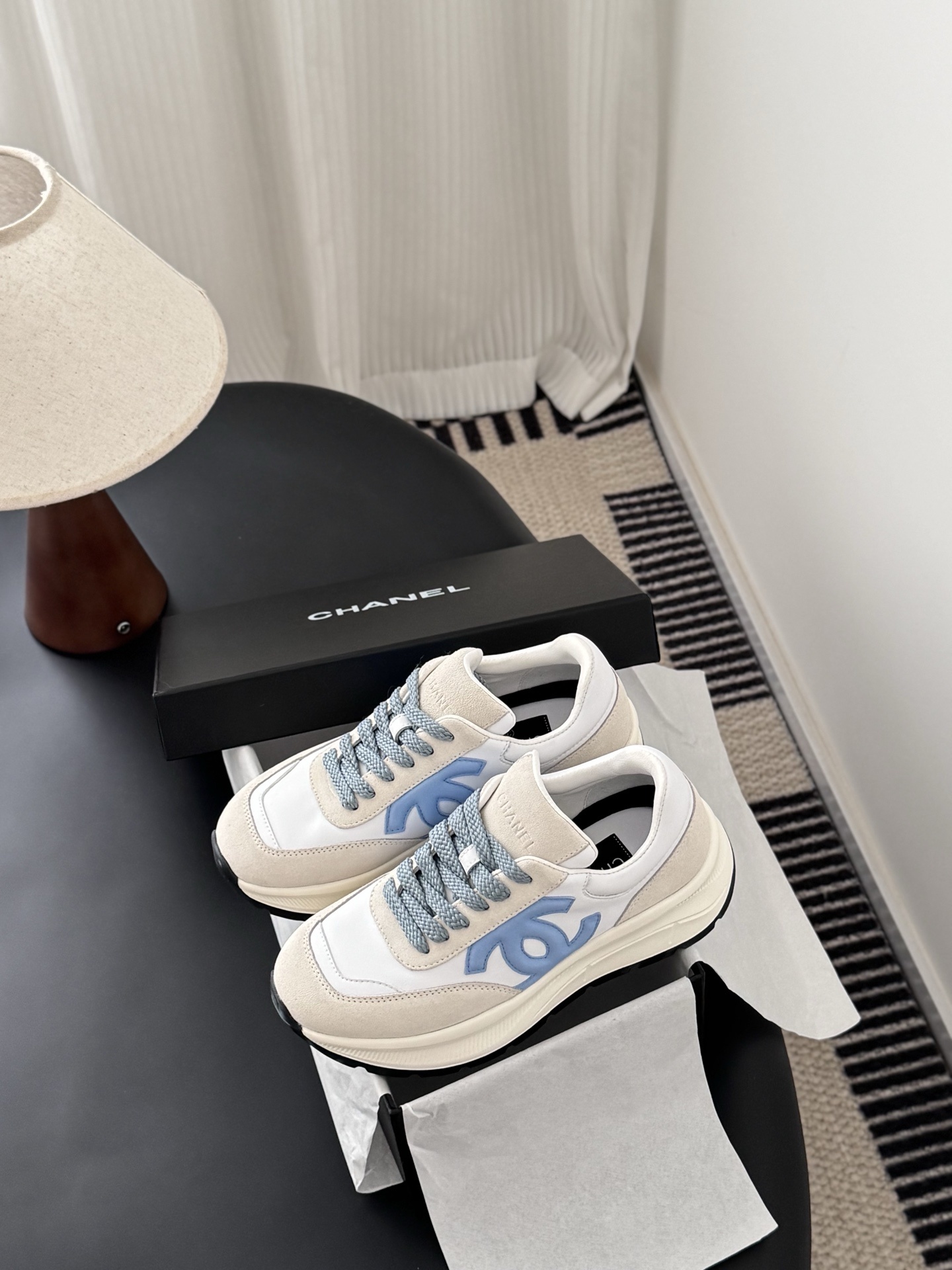 A pair of Chanel white and beige sneakers with light blue CC logo and laces next to a black Chanel shoe box. - Hình 9