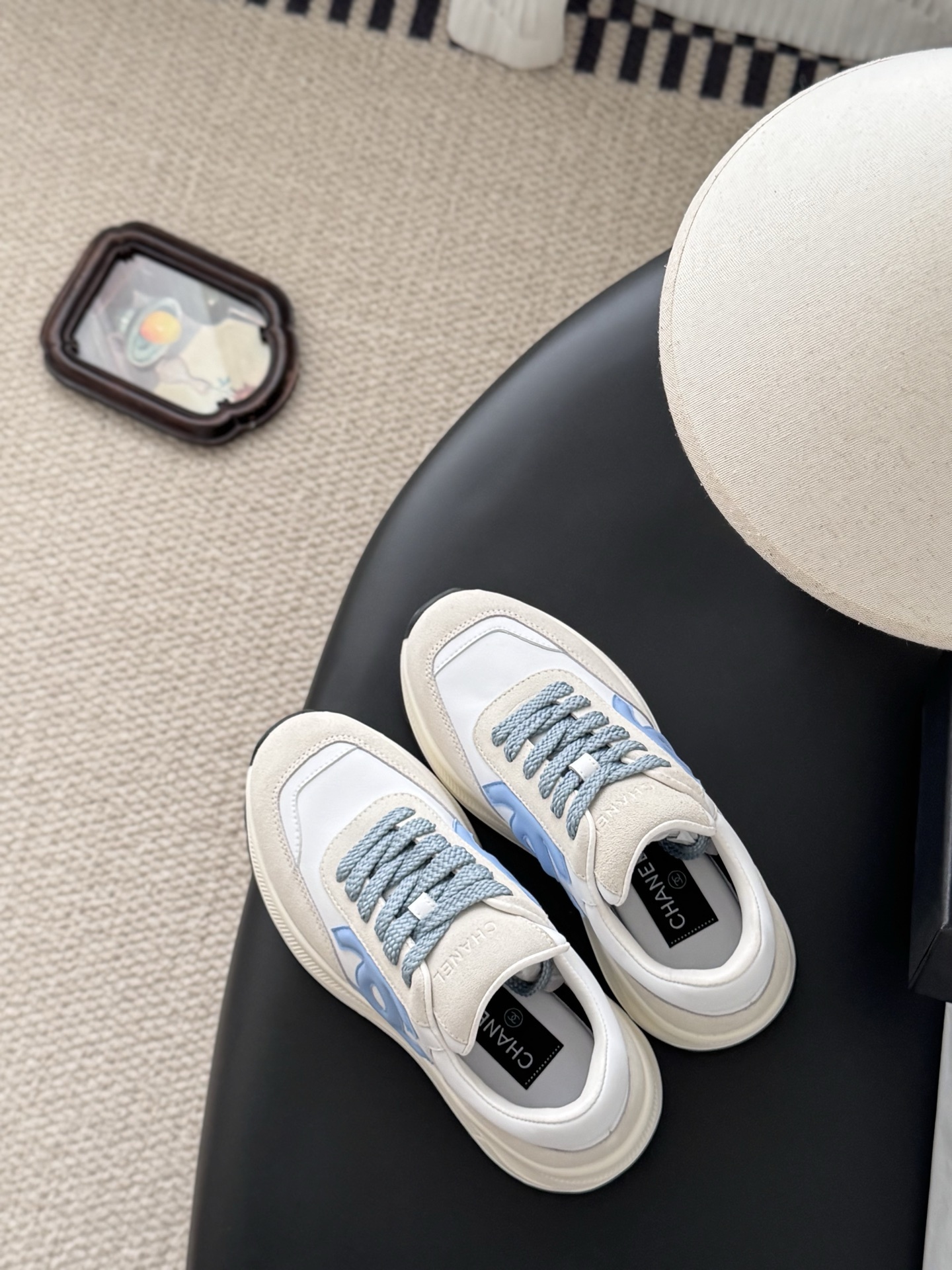 A pair of Chanel white and beige sneakers with light blue CC logo and laces next to a black Chanel shoe box. - Hình 8