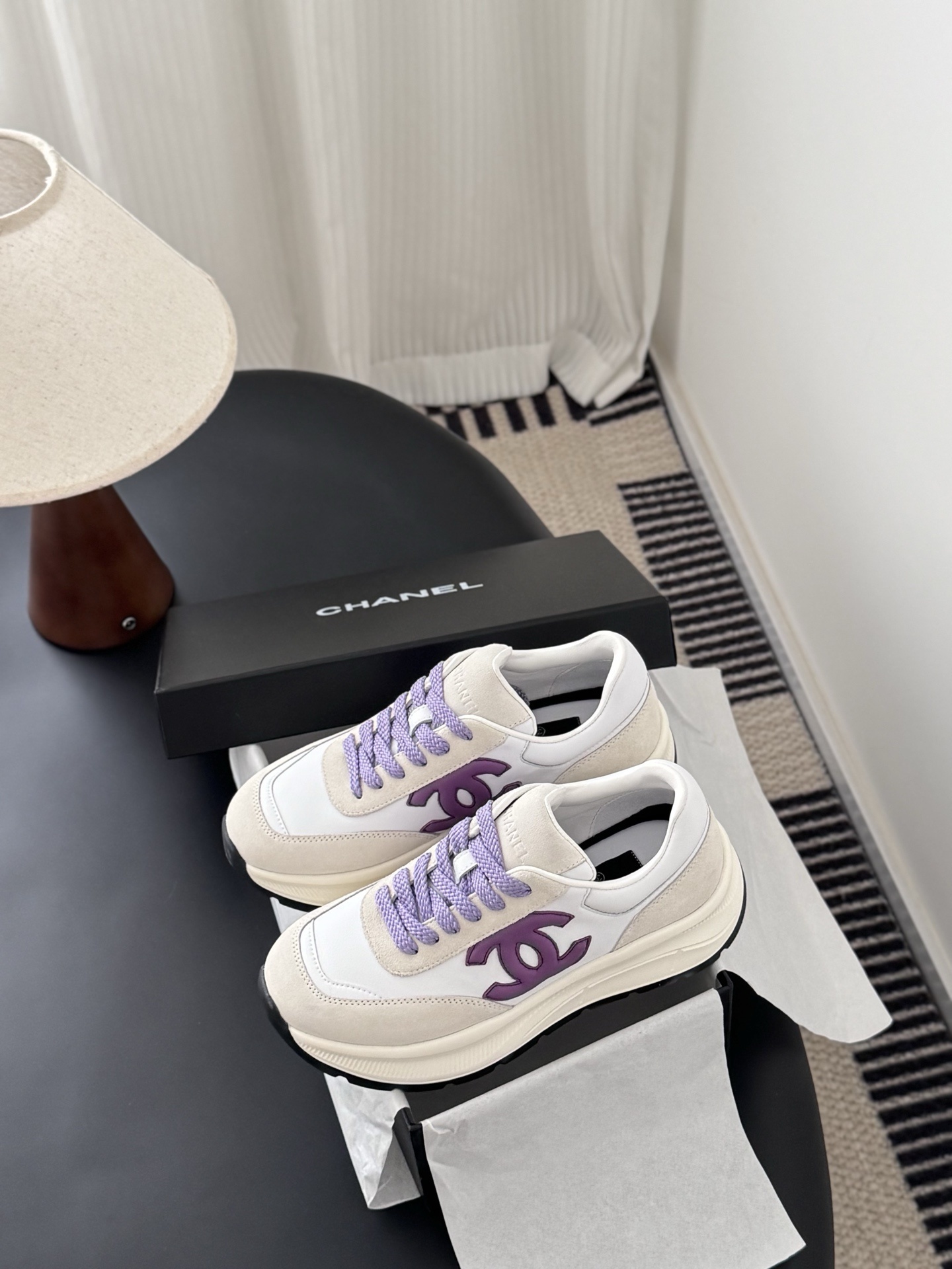 A pair of white and purple Chanel suede leather sneakers displayed next to their original black Chanel box on a black surface. - Hình 9