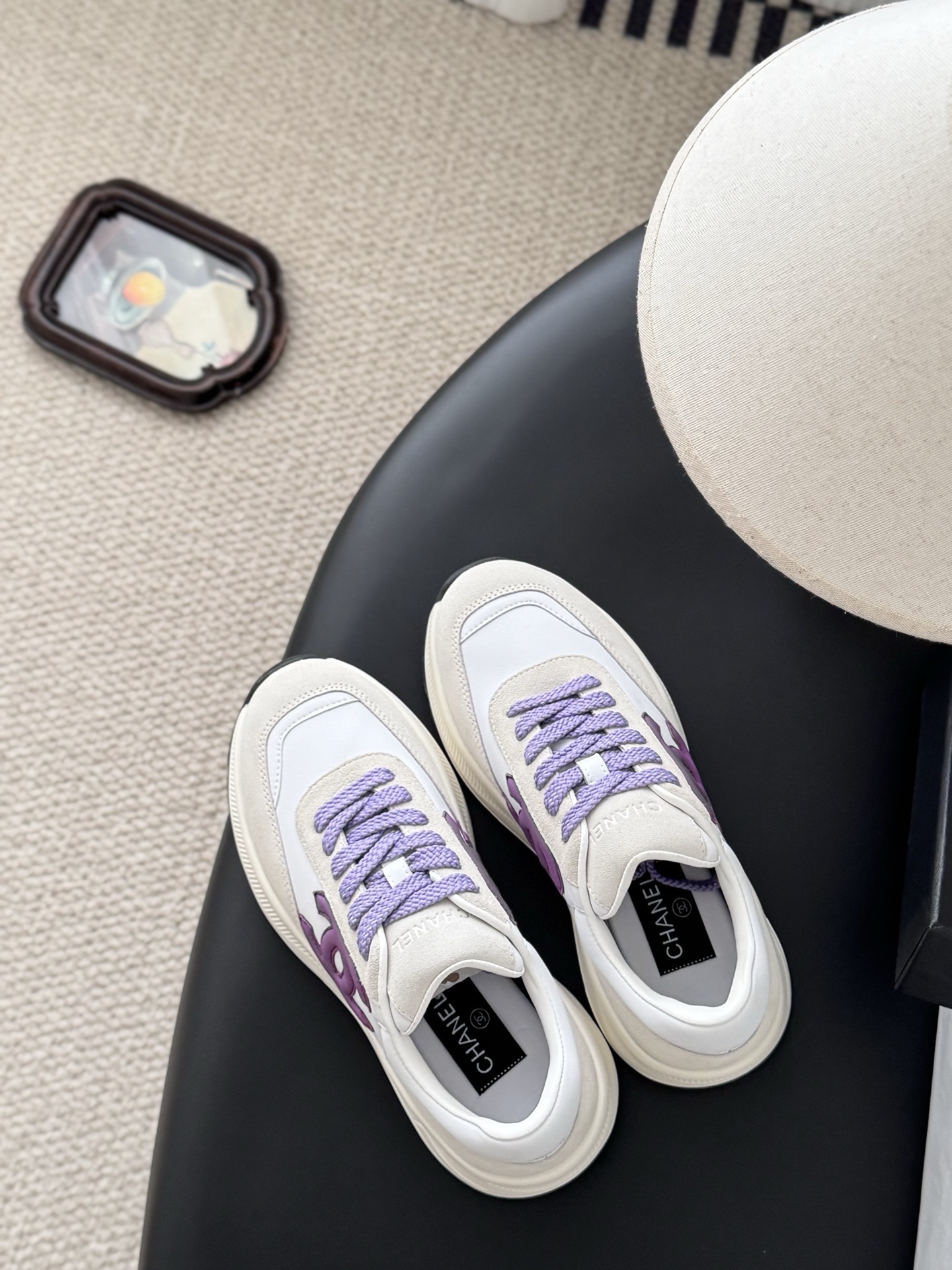 A pair of white and purple Chanel suede leather sneakers displayed next to their original black Chanel box on a black surface. - Hình 8