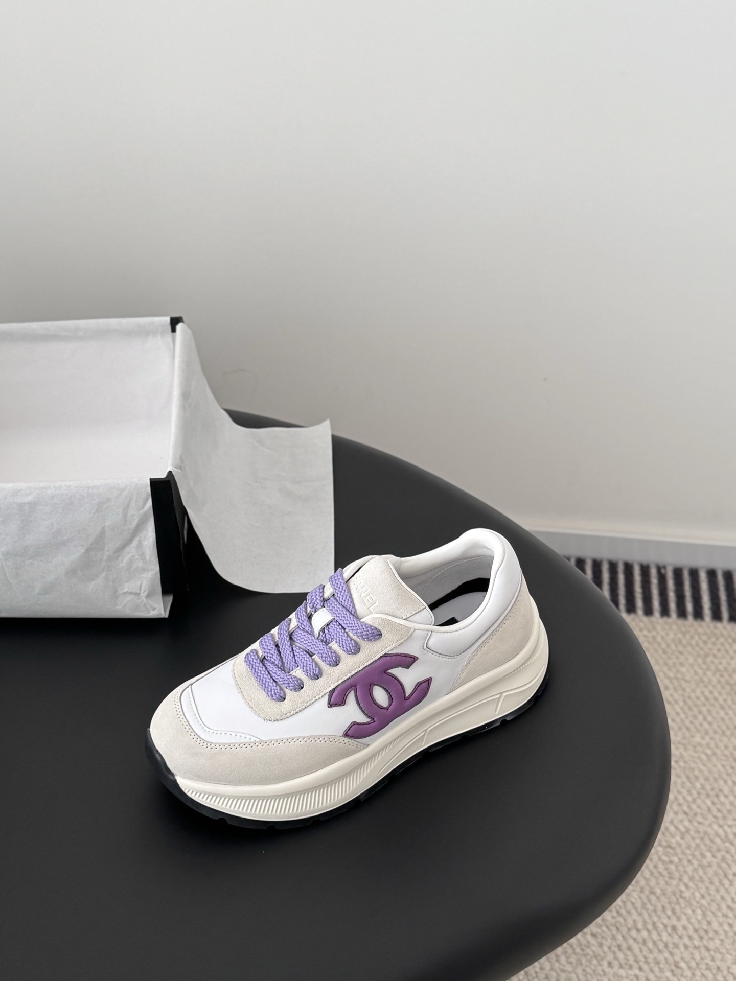 A pair of white and purple Chanel suede leather sneakers displayed next to their original black Chanel box on a black surface. - Hình 6