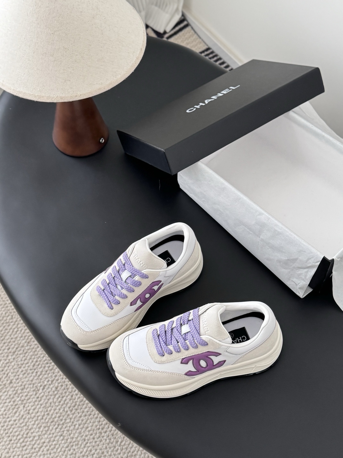 A pair of white and purple Chanel suede leather sneakers displayed next to their original black Chanel box on a black surface. - Hình 5