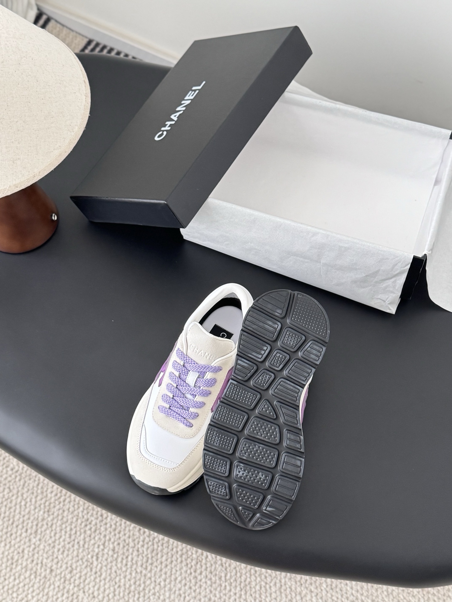 A pair of white and purple Chanel suede leather sneakers displayed next to their original black Chanel box on a black surface. - Hình 4