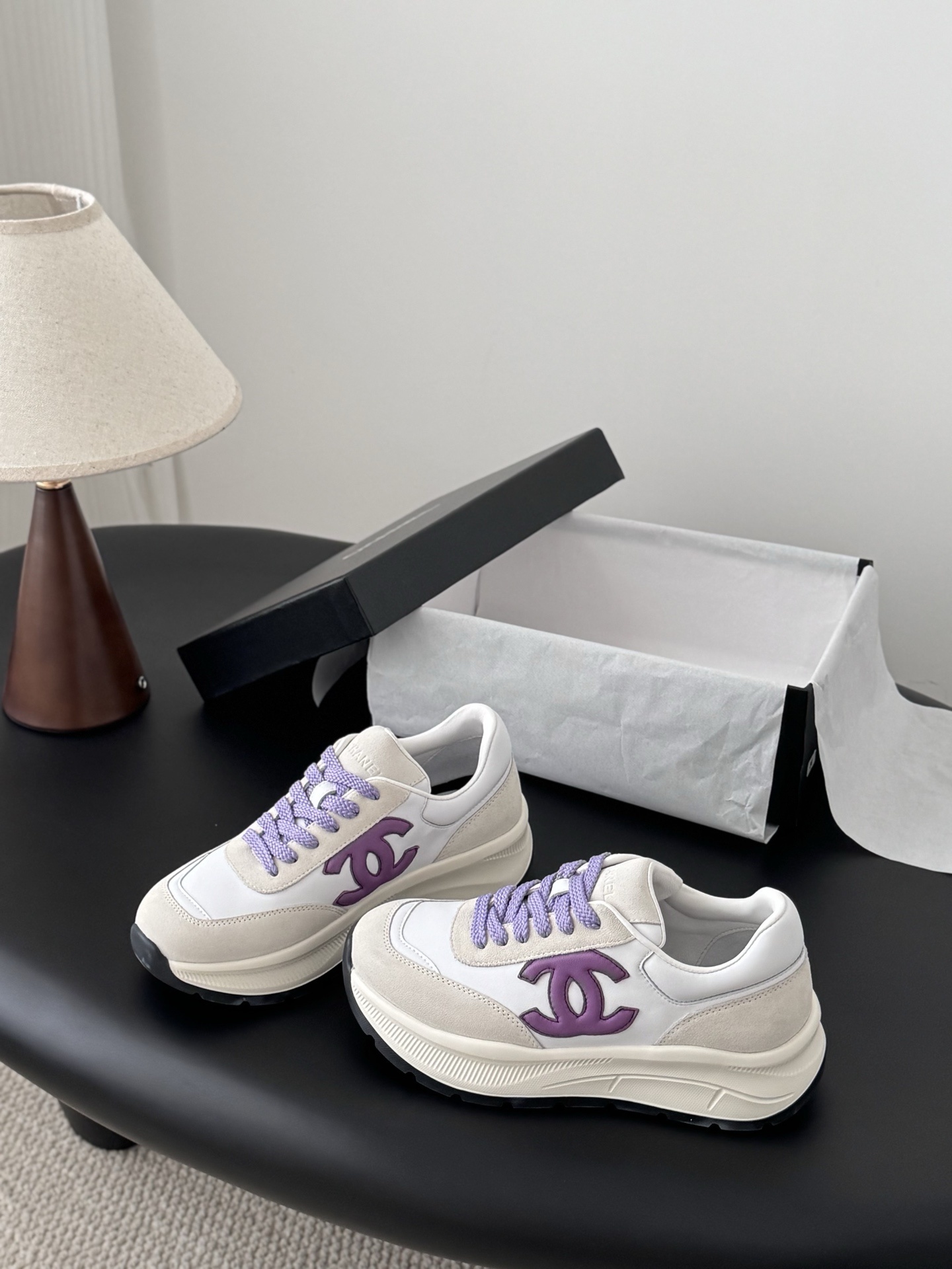 A pair of white and purple Chanel suede leather sneakers displayed next to their original black Chanel box on a black surface. - Hình 2