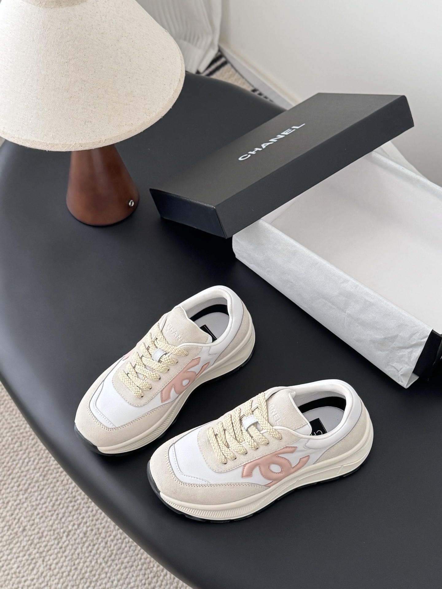 A pair of Chanel women's sneakers in white and beige suede with a pink CC logo, displayed next to a black Chanel shoe box. - Hình 5