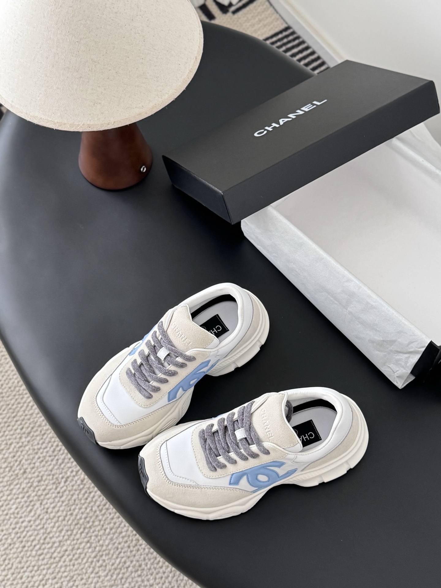 A pair of Chanel white and light blue leather sneakers with grey laces placed on a black surface next to an open Chanel shoe box. - Hình 5