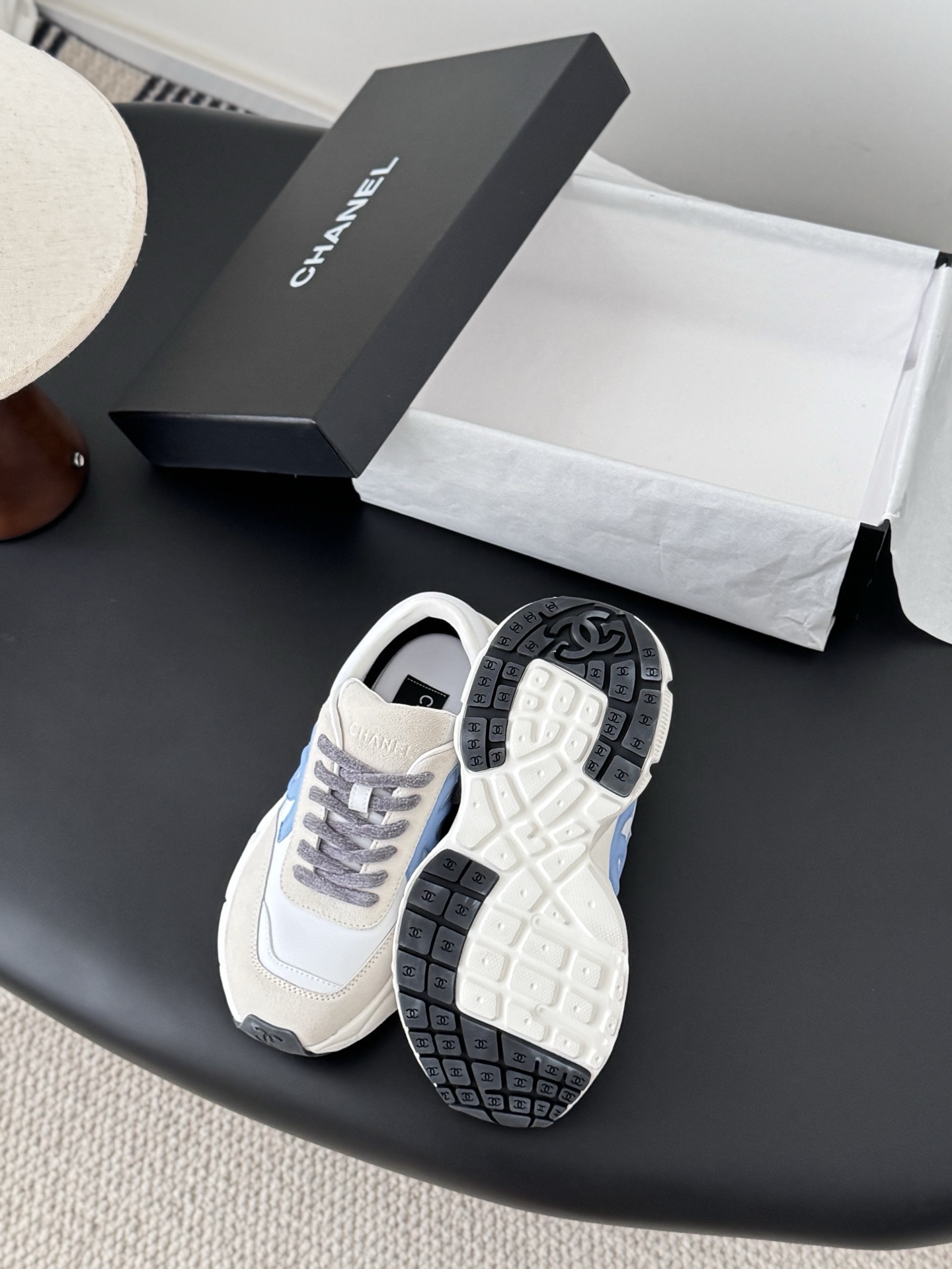 A pair of Chanel white and light blue leather sneakers with grey laces placed on a black surface next to an open Chanel shoe box. - Hình 4