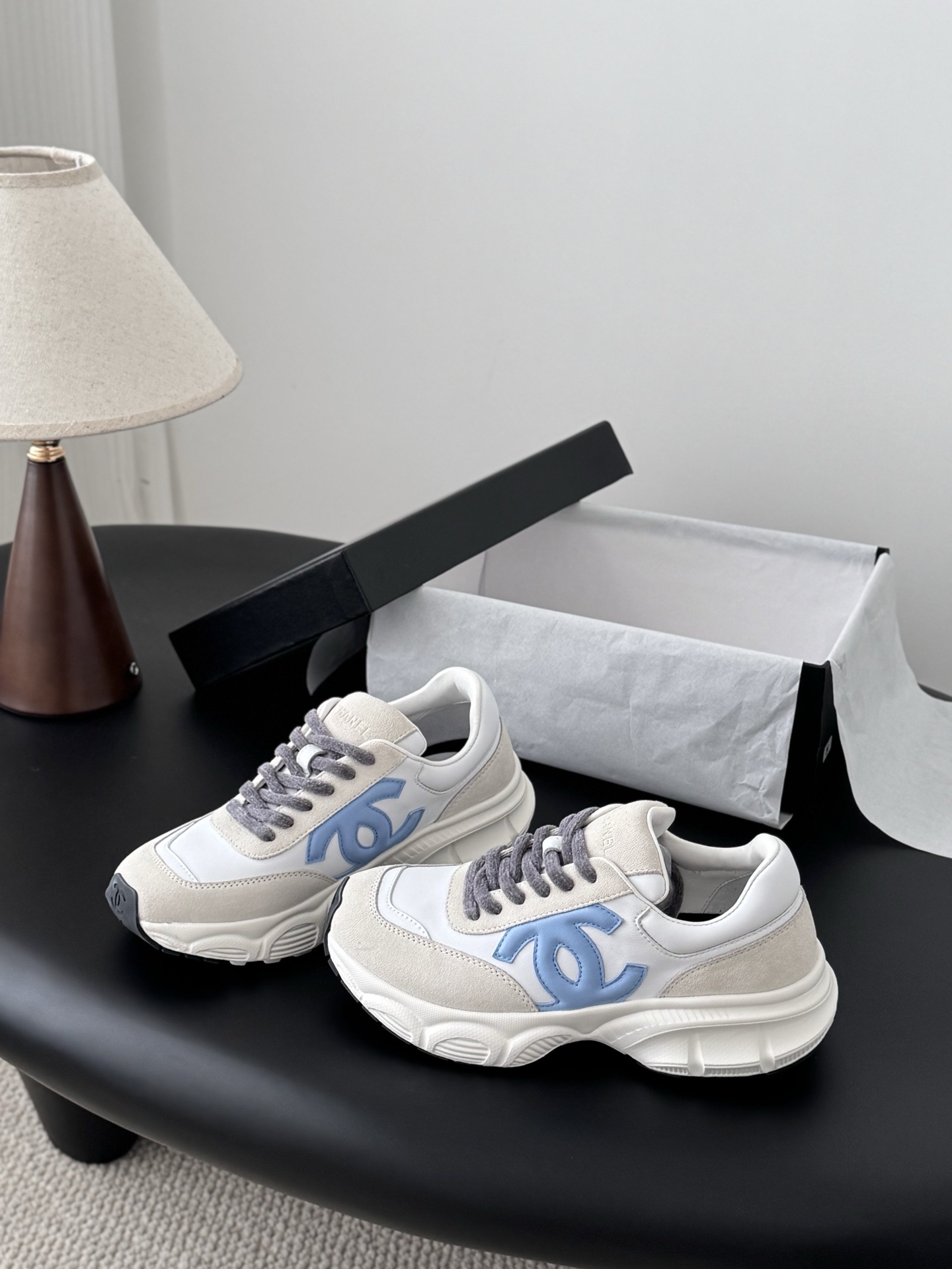 A pair of Chanel white and light blue leather sneakers with grey laces placed on a black surface next to an open Chanel shoe box. - Hình 2