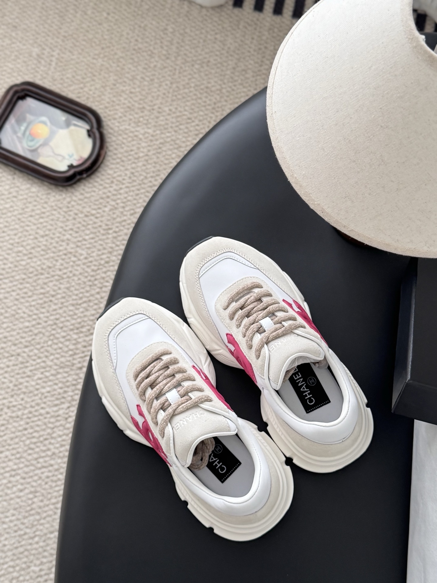 A pair of white and beige Chanel sneakers with a prominent pink CC logo on the side, sitting next to a black Chanel box. - Hình 8