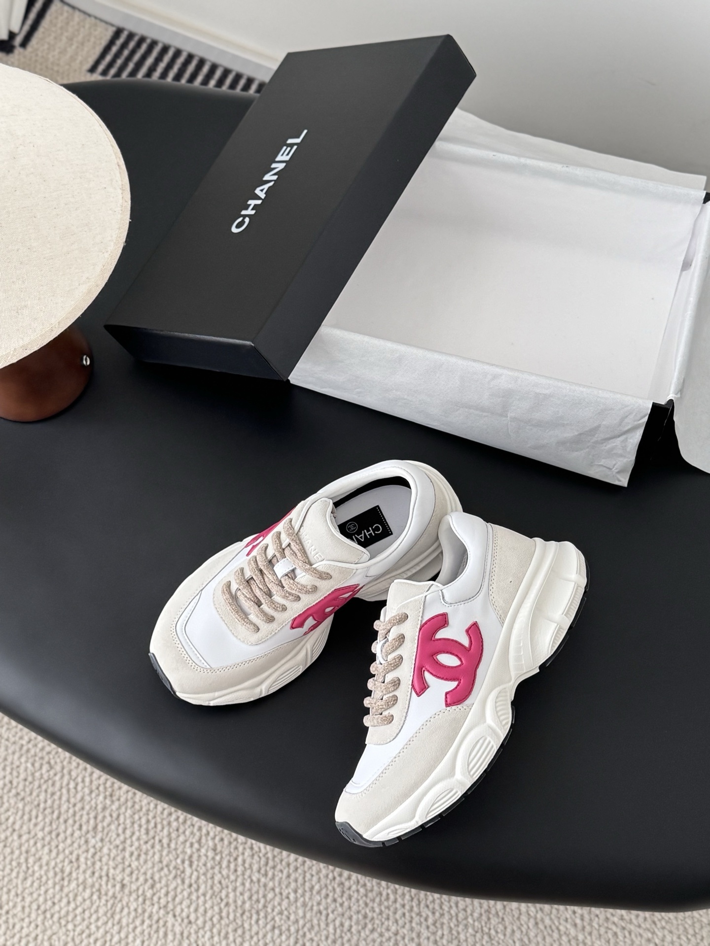 A pair of white and beige Chanel sneakers with a prominent pink CC logo on the side, sitting next to a black Chanel box. - Hình 3