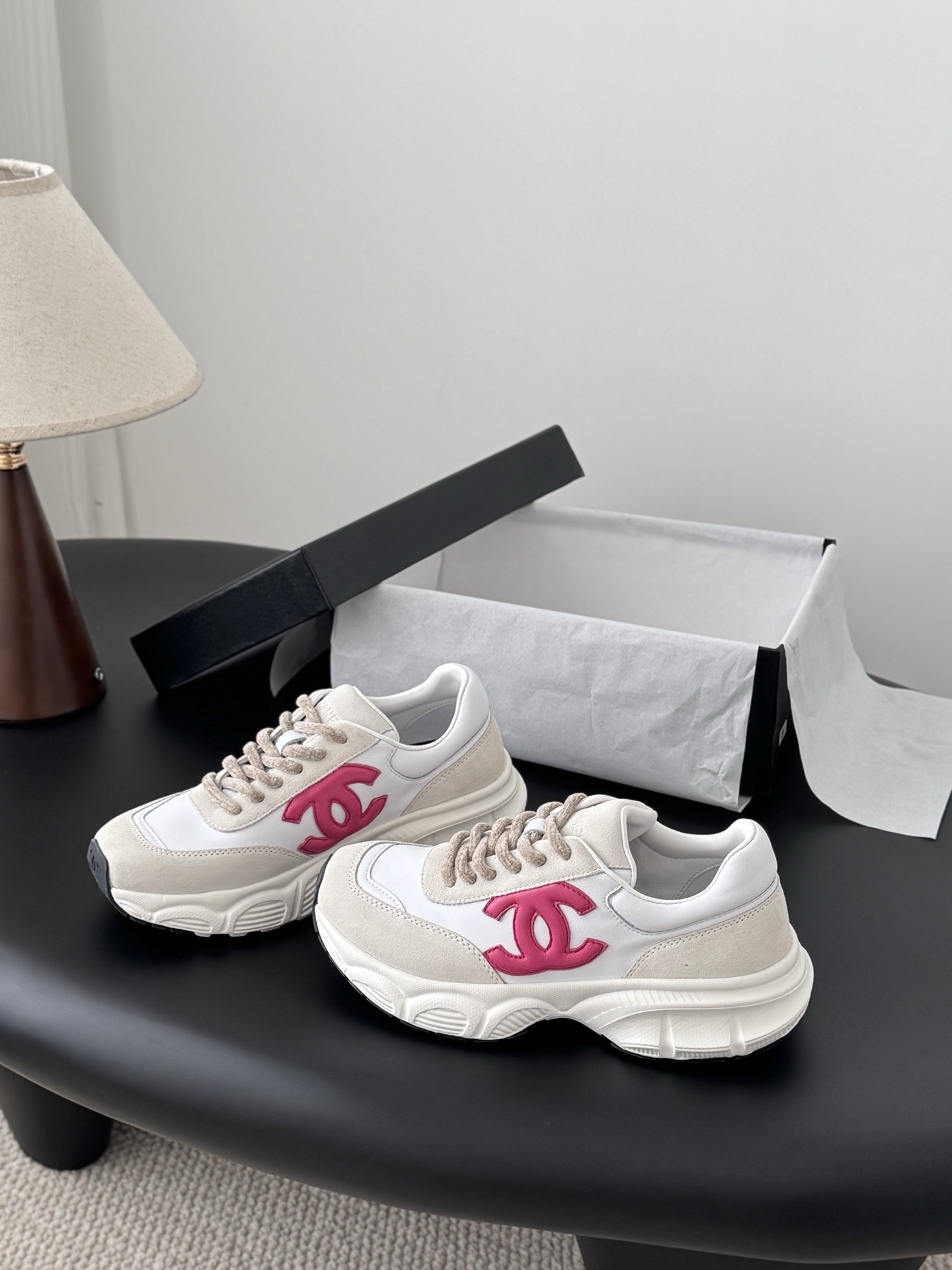 A pair of white and beige Chanel sneakers with a prominent pink CC logo on the side, sitting next to a black Chanel box. - Hình 2