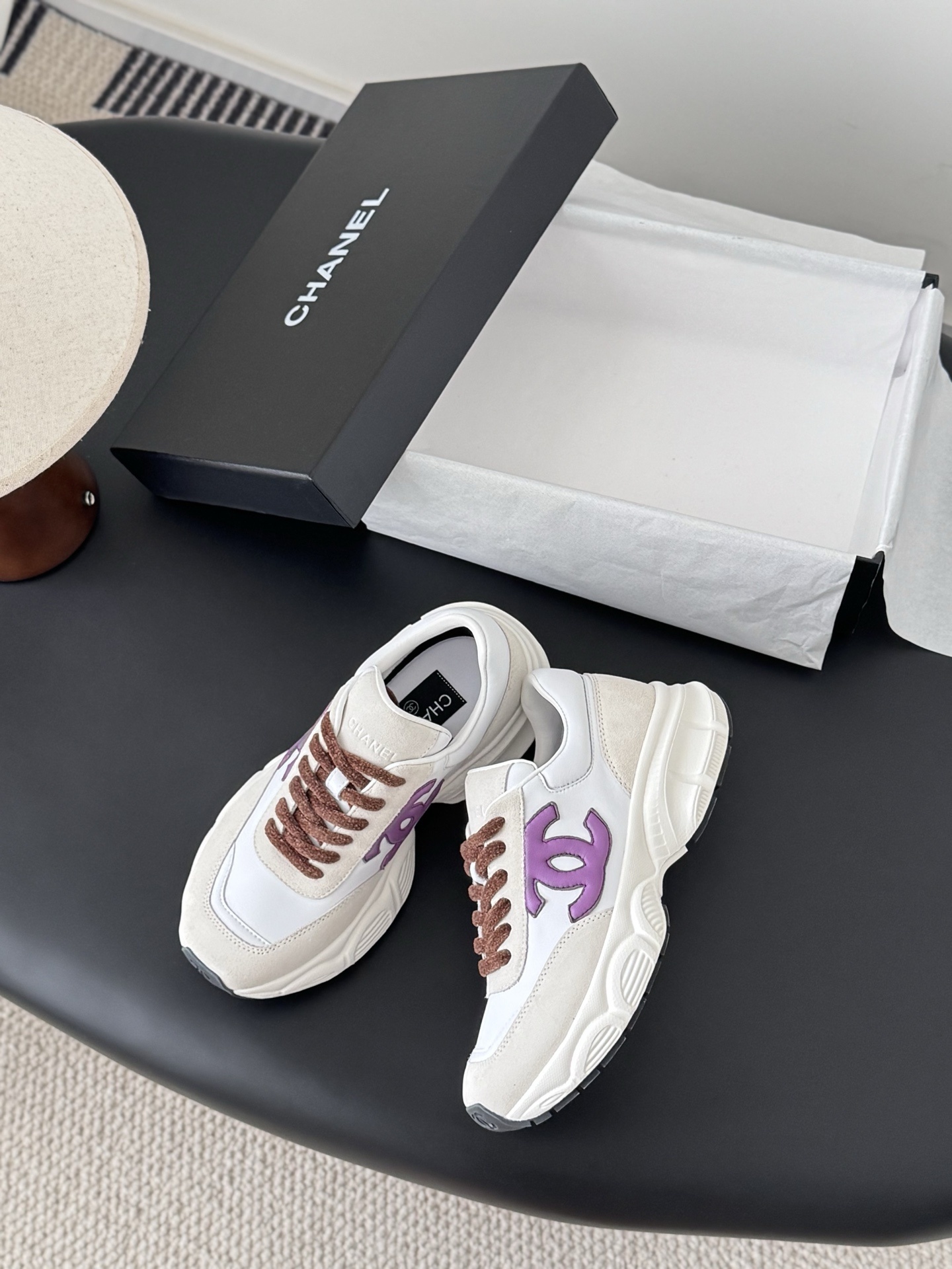 A pair of white and cream Chanel sneakers with purple CC logos and brown laces next to their original box. - Hình 3