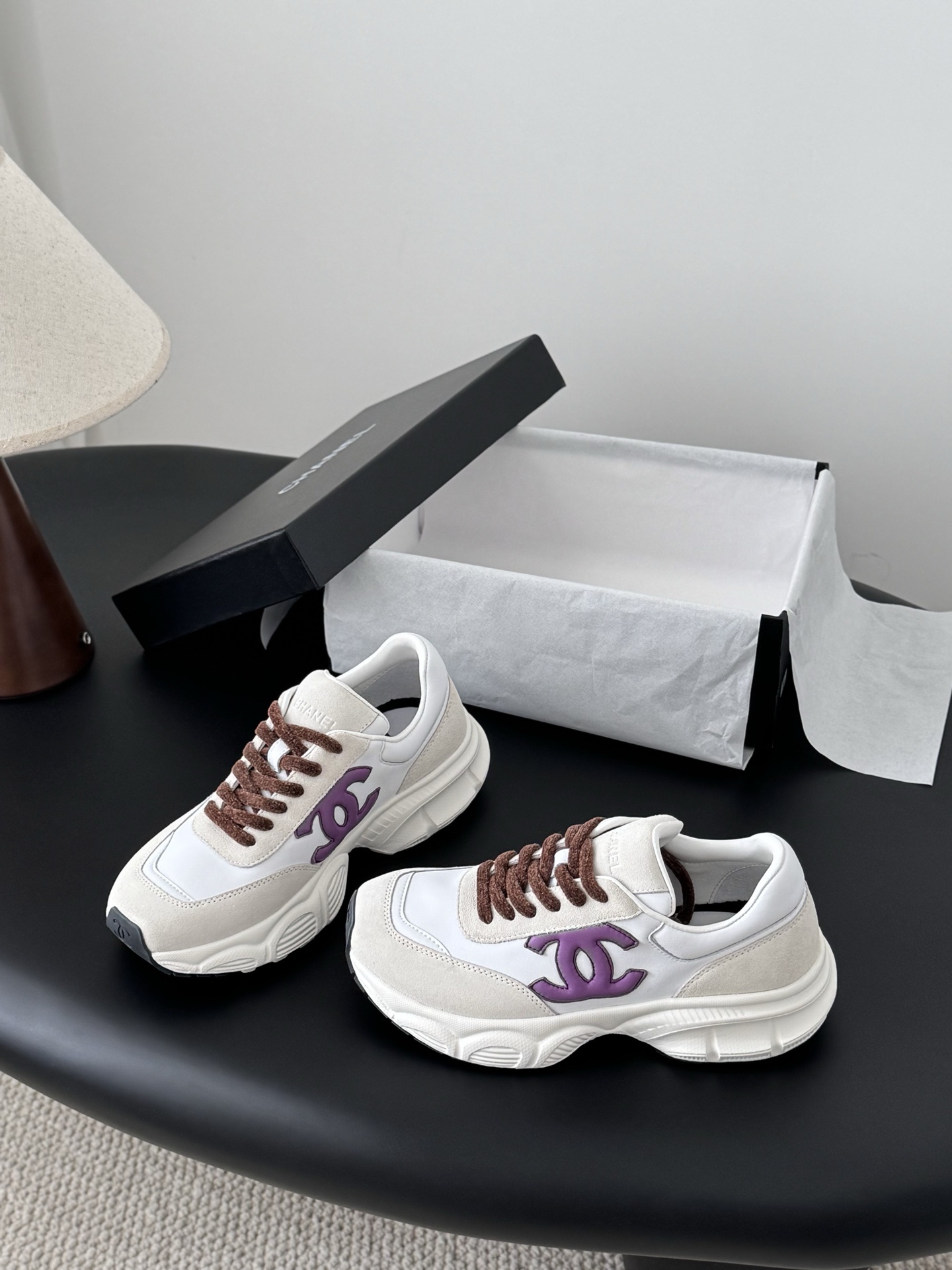 A pair of white and cream Chanel sneakers with purple CC logos and brown laces next to their original box. - Hình 2