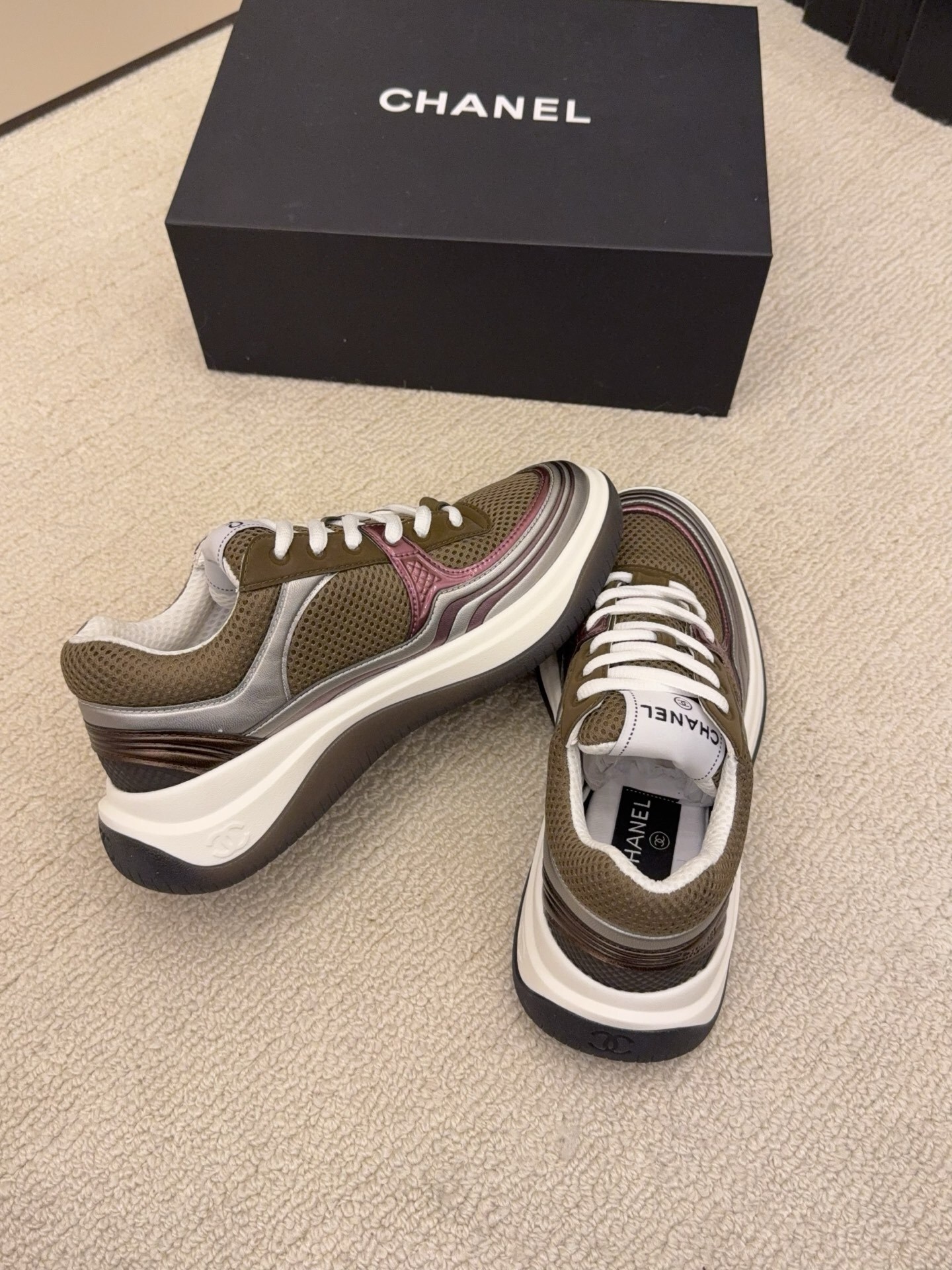 Two pairs of Chanel luxury sneakers in metallic olive and burgundy colorways displayed on a black box and carpet. - Hình 9