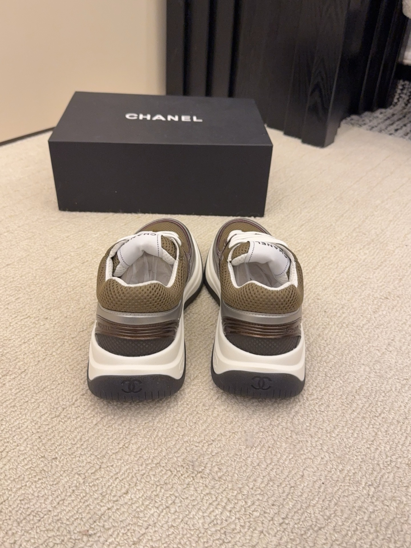 Two pairs of Chanel luxury sneakers in metallic olive and burgundy colorways displayed on a black box and carpet. - Hình 8