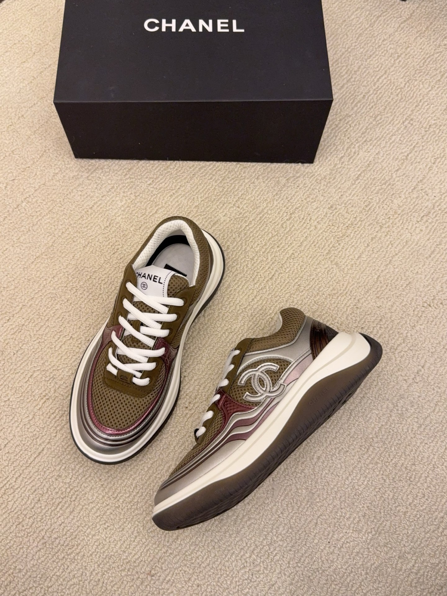 Two pairs of Chanel luxury sneakers in metallic olive and burgundy colorways displayed on a black box and carpet. - Hình 7