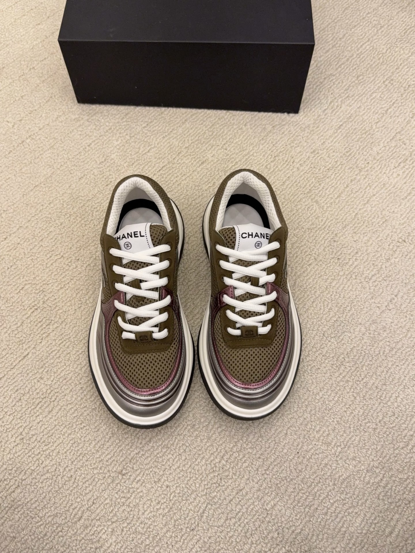 Two pairs of Chanel luxury sneakers in metallic olive and burgundy colorways displayed on a black box and carpet. - Hình 6