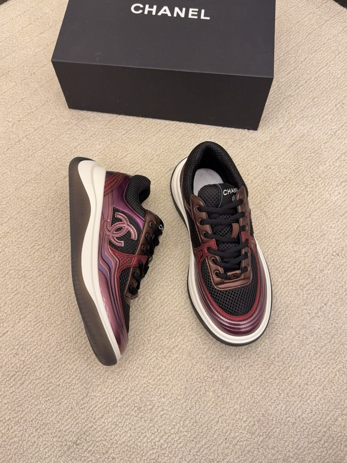 Two pairs of Chanel luxury sneakers in metallic olive and burgundy colorways displayed on a black box and carpet. - Hình 3