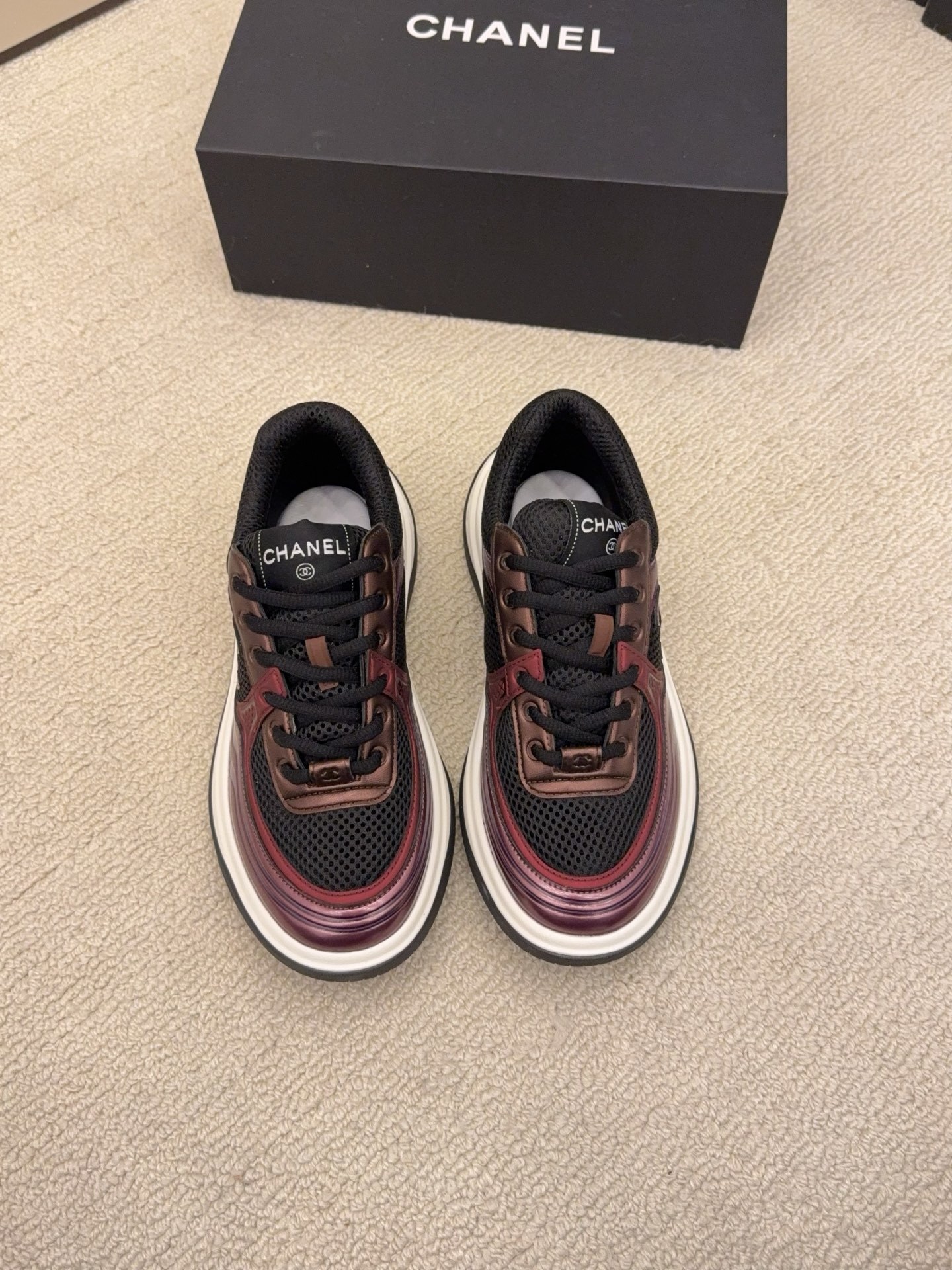 Two pairs of Chanel luxury sneakers in metallic olive and burgundy colorways displayed on a black box and carpet. - Hình 2