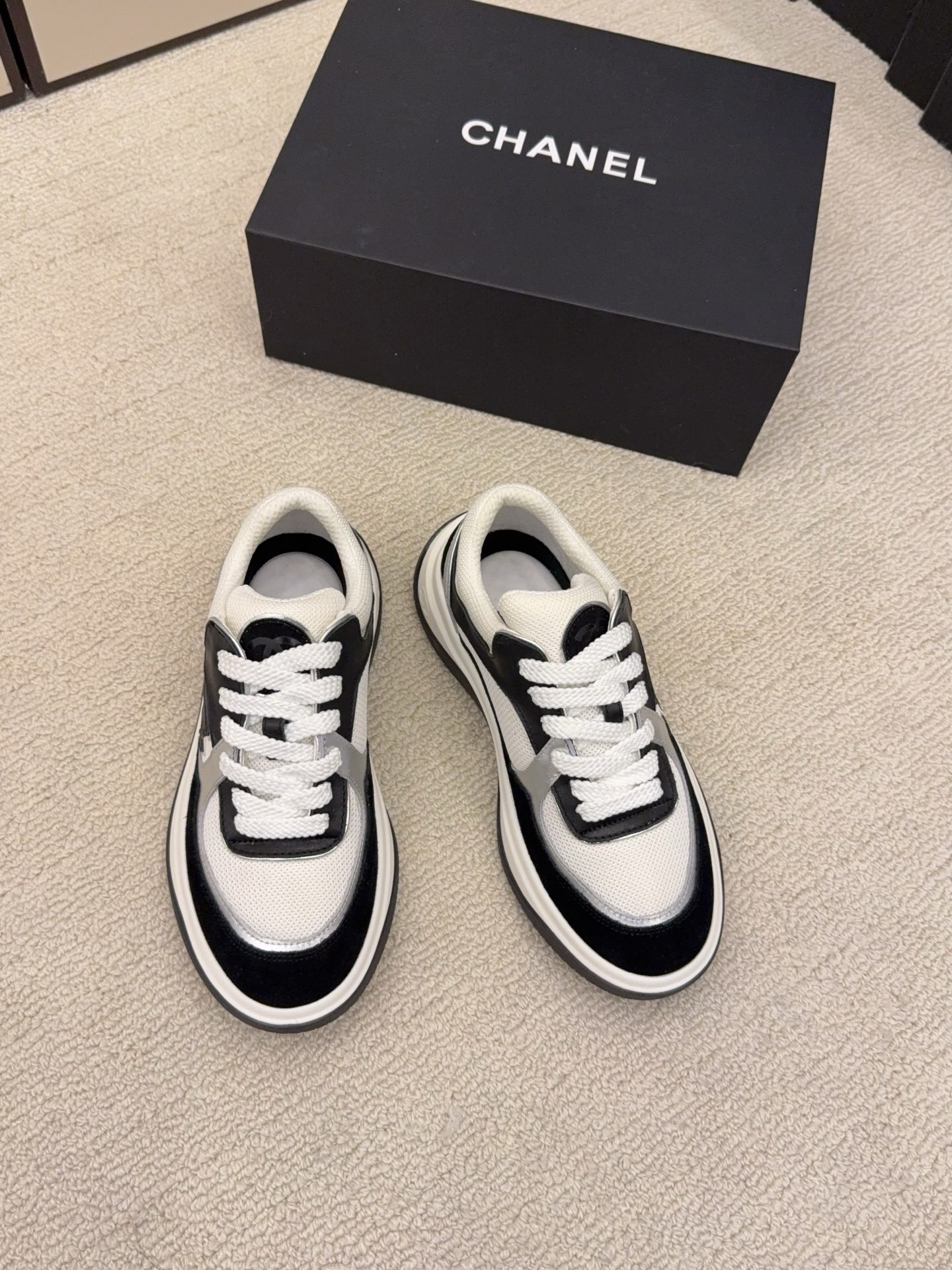 Top view of a pair of Chanel black and white sneakers with the iconic CC logo next to a black Chanel box - Hình 2
