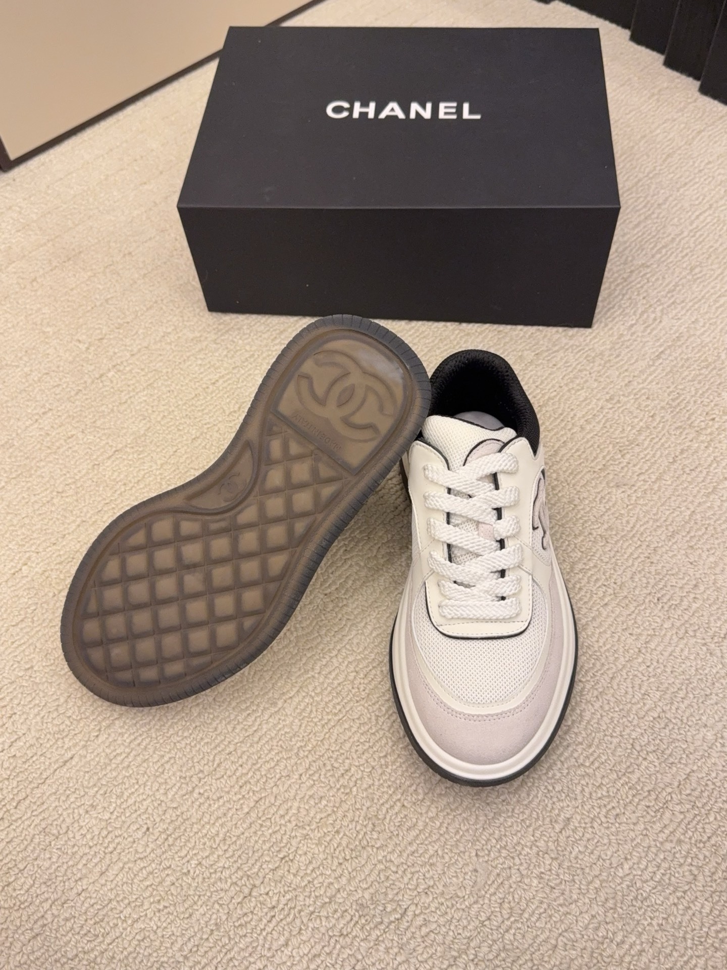 Pair of Chanel low-top sneakers in beige and black with the interlocking CC logo on the side next to a black shoe box. - Hình 9