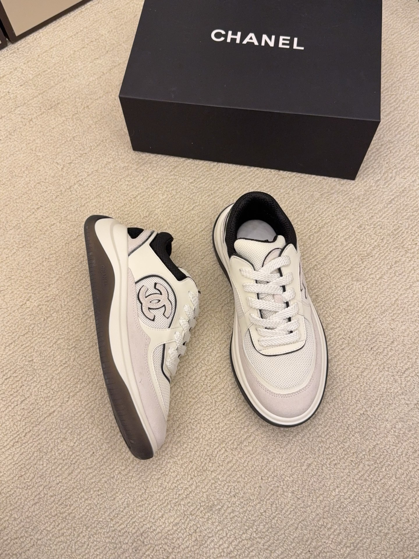 Pair of Chanel low-top sneakers in beige and black with the interlocking CC logo on the side next to a black shoe box. - Hình 7