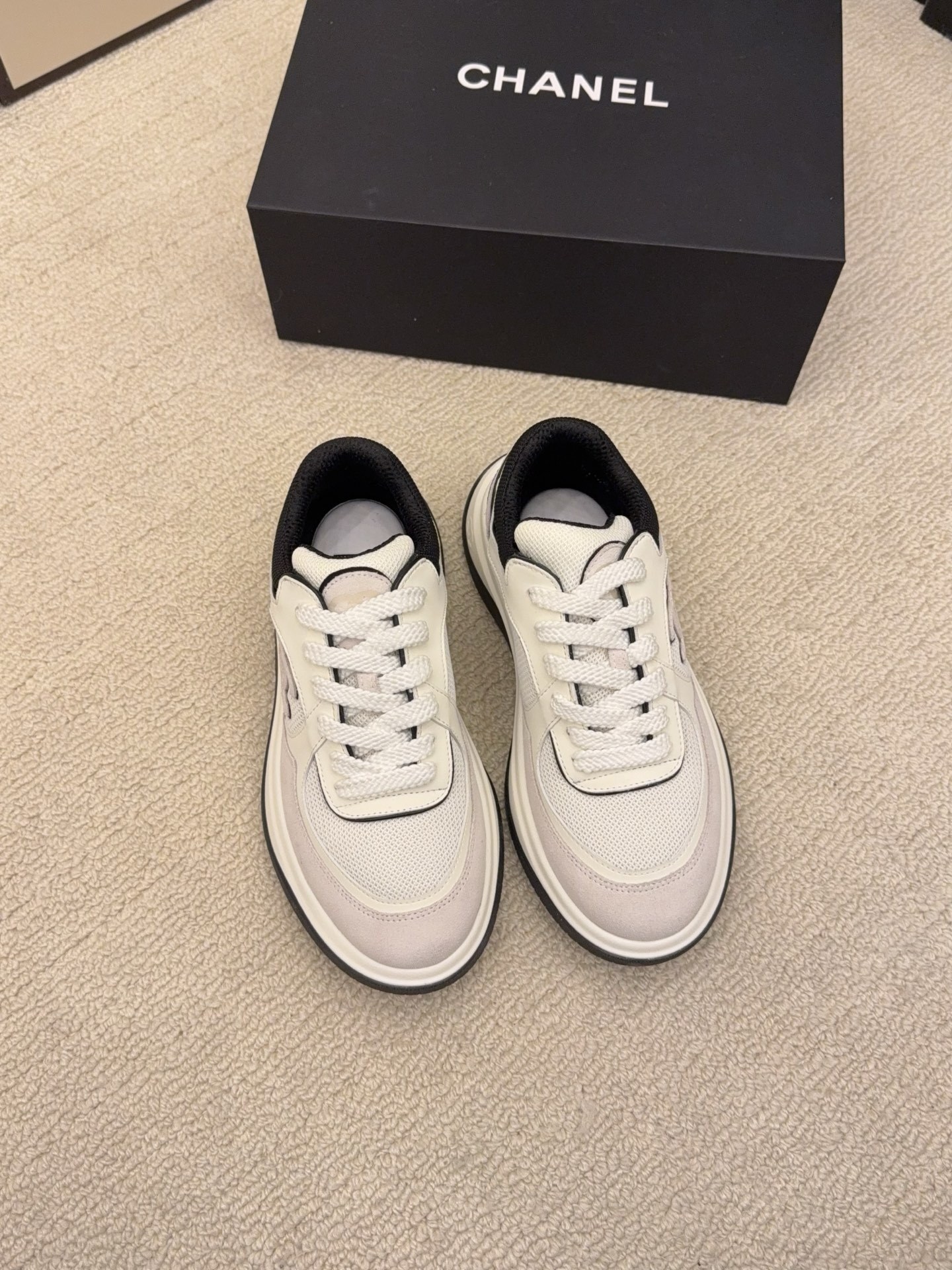 Pair of Chanel low-top sneakers in beige and black with the interlocking CC logo on the side next to a black shoe box. - Hình 6