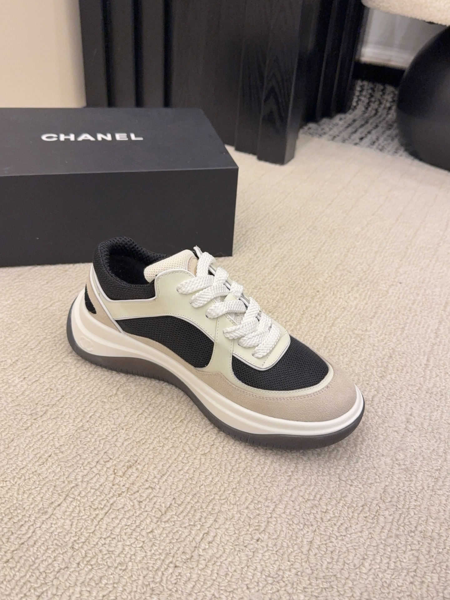 Pair of Chanel low-top sneakers in beige and black with the interlocking CC logo on the side next to a black shoe box. - Hình 5
