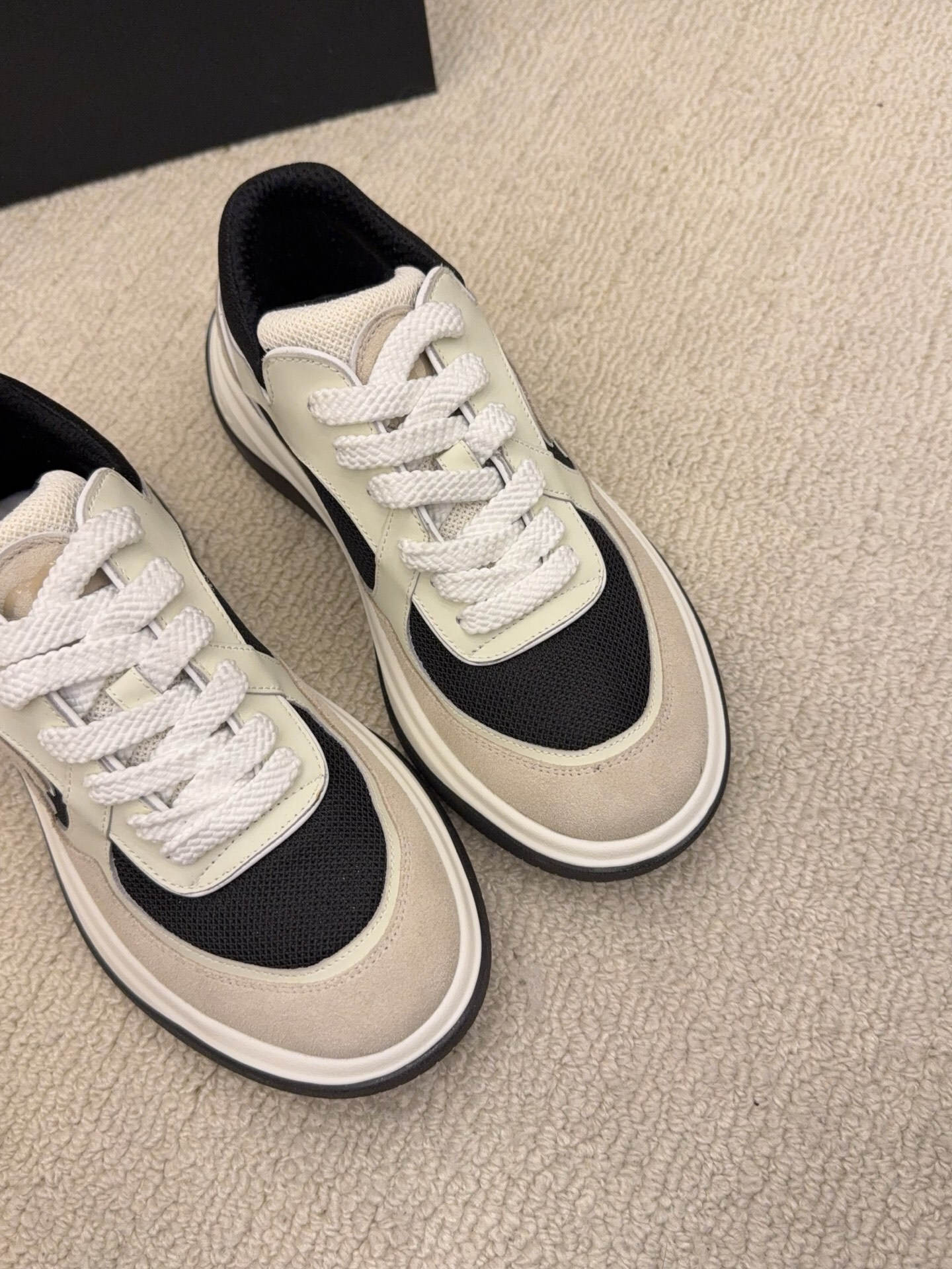 Pair of Chanel low-top sneakers in beige and black with the interlocking CC logo on the side next to a black shoe box. - Hình 4