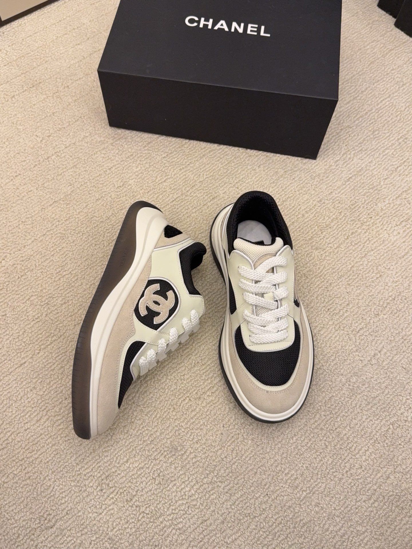Pair of Chanel low-top sneakers in beige and black with the interlocking CC logo on the side next to a black shoe box. - Hình 3