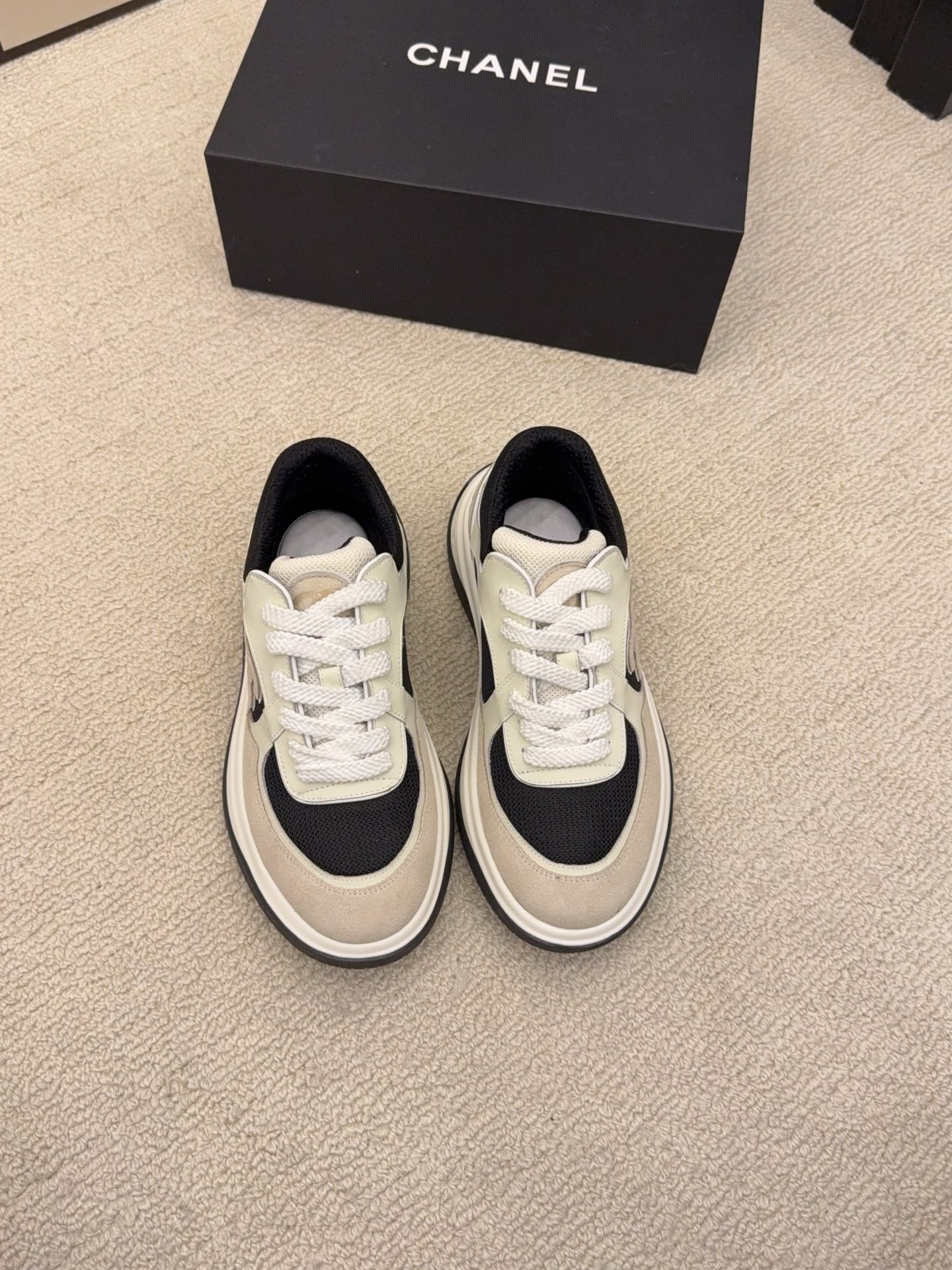 Pair of Chanel low-top sneakers in beige and black with the interlocking CC logo on the side next to a black shoe box. - Hình 2