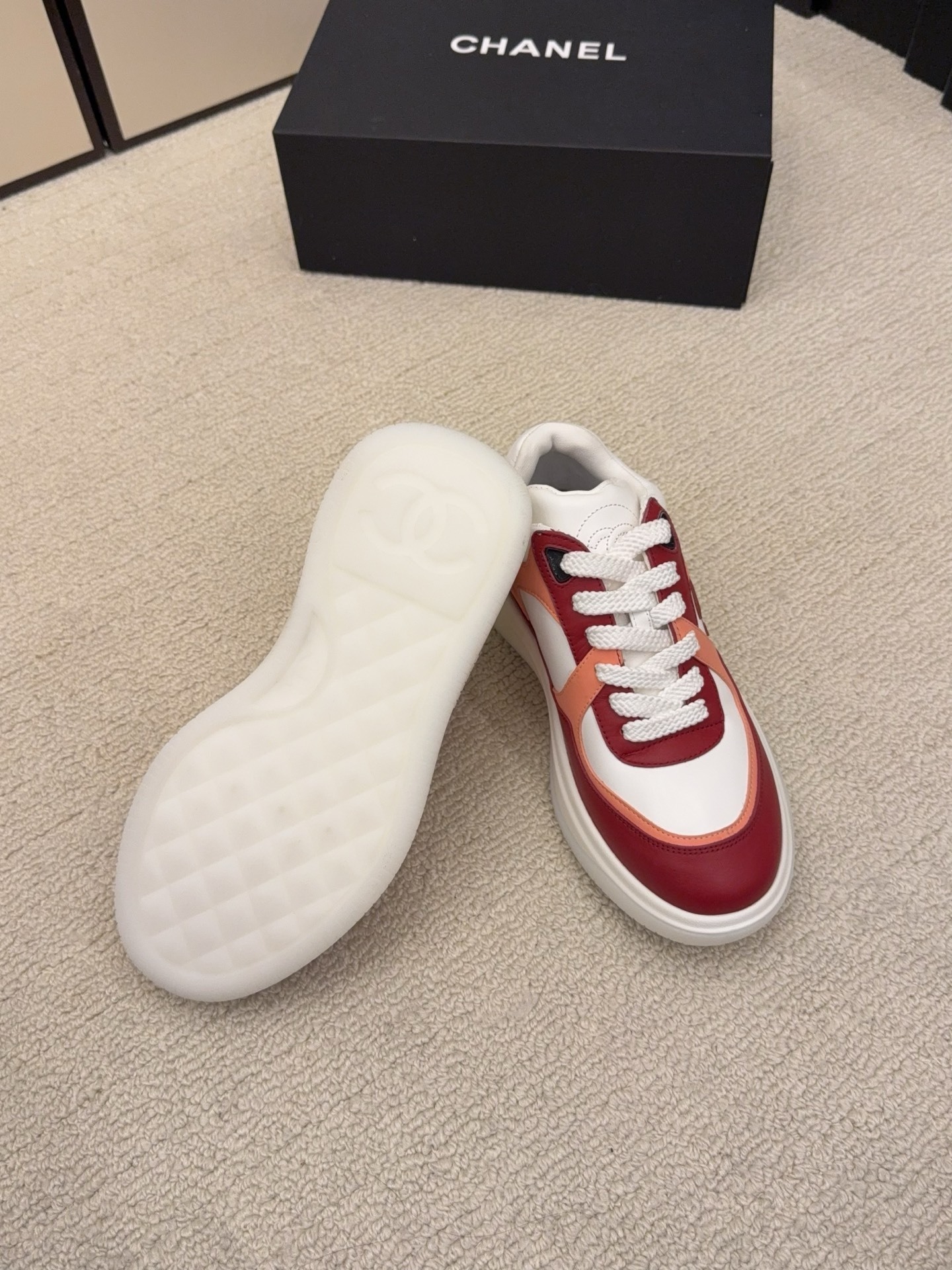 A pair of Chanel women's low-top sneakers in red, white, and orange leather with thick white laces next to a black Chanel box. - Hình 9