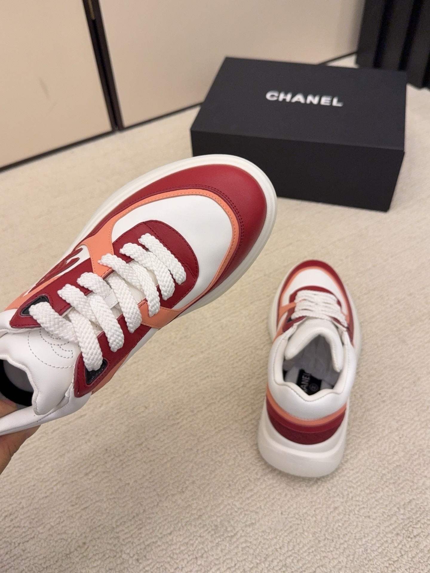 A pair of Chanel women's low-top sneakers in red, white, and orange leather with thick white laces next to a black Chanel box. - Hình 8