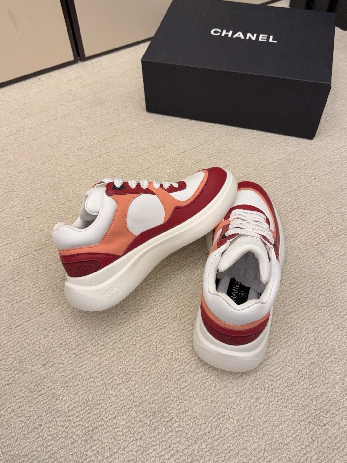 A pair of Chanel women's low-top sneakers in red, white, and orange leather with thick white laces next to a black Chanel box. - Hình 7