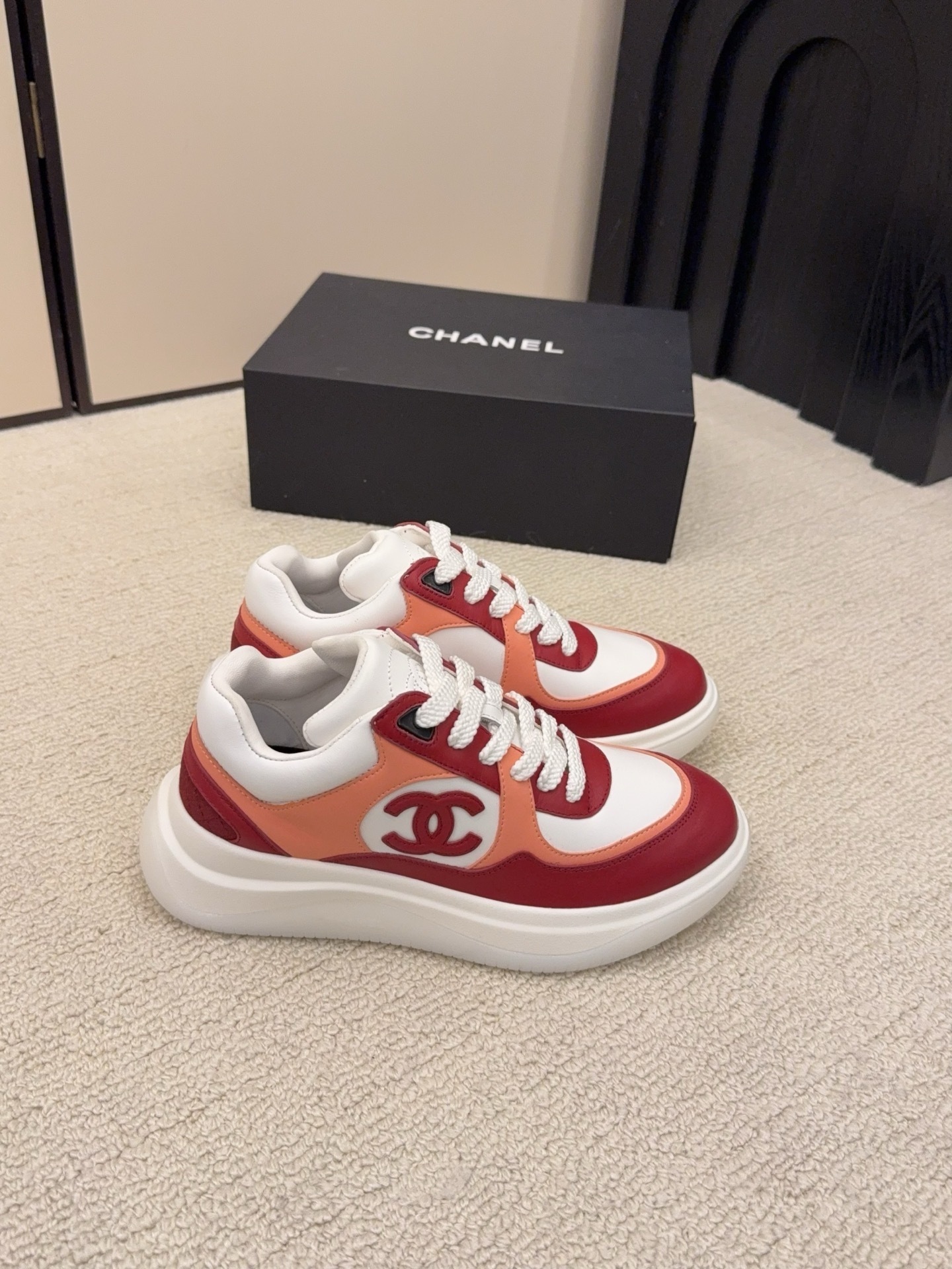 A pair of Chanel women's low-top sneakers in red, white, and orange leather with thick white laces next to a black Chanel box. - Hình 5
