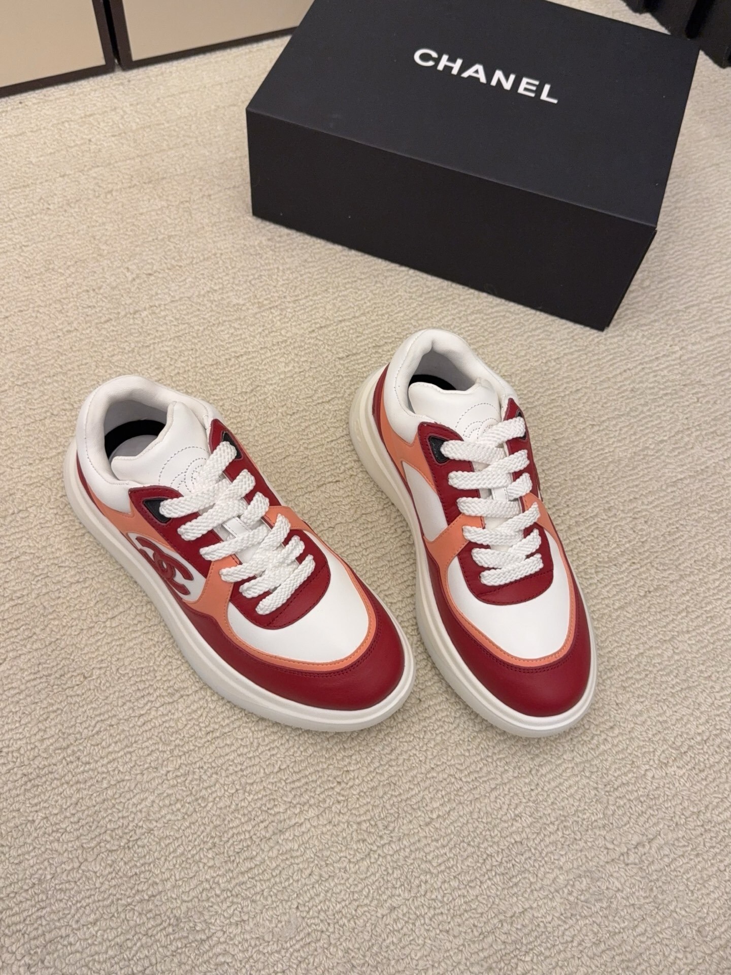 A pair of Chanel women's low-top sneakers in red, white, and orange leather with thick white laces next to a black Chanel box. - Hình 3