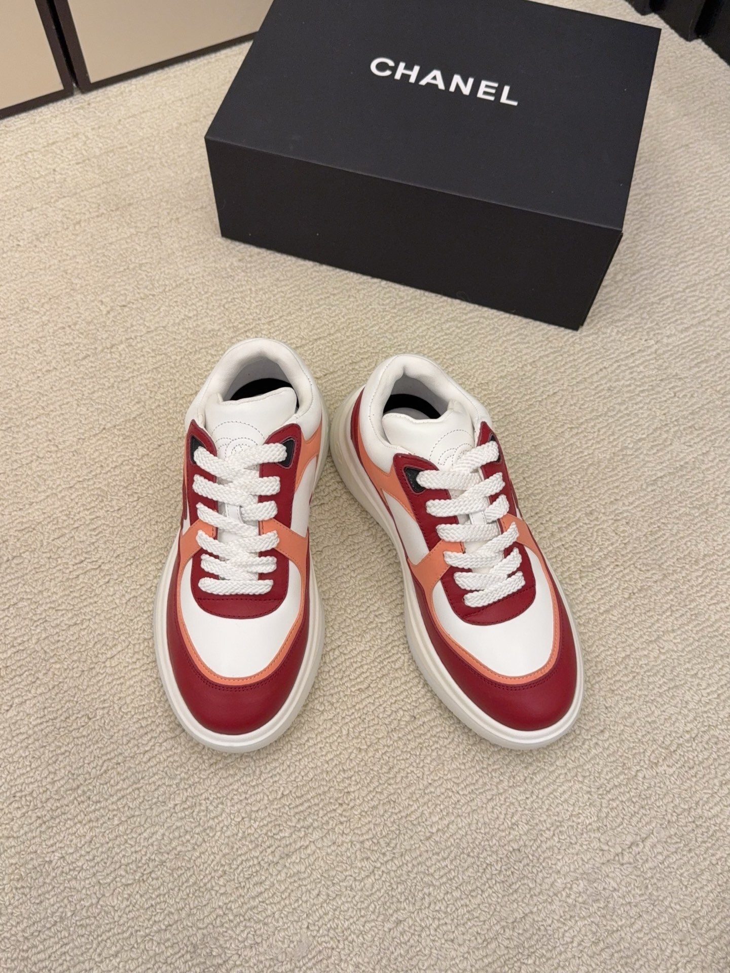 A pair of Chanel women's low-top sneakers in red, white, and orange leather with thick white laces next to a black Chanel box. - Hình 2