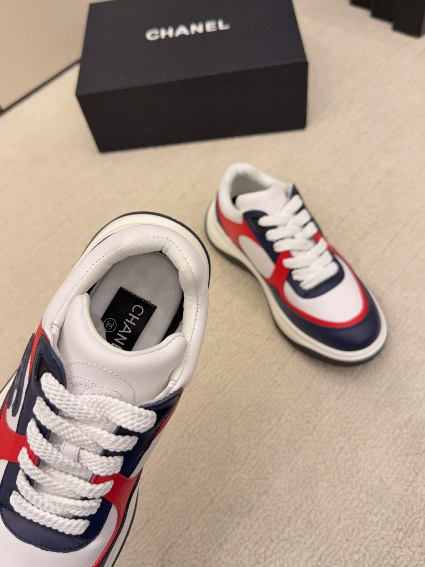 A pair of Chanel tricolor sneakers in white, navy blue, and red with a chunky sole and the CC logo, displayed next to a black Chanel shoe box. - Hình 9