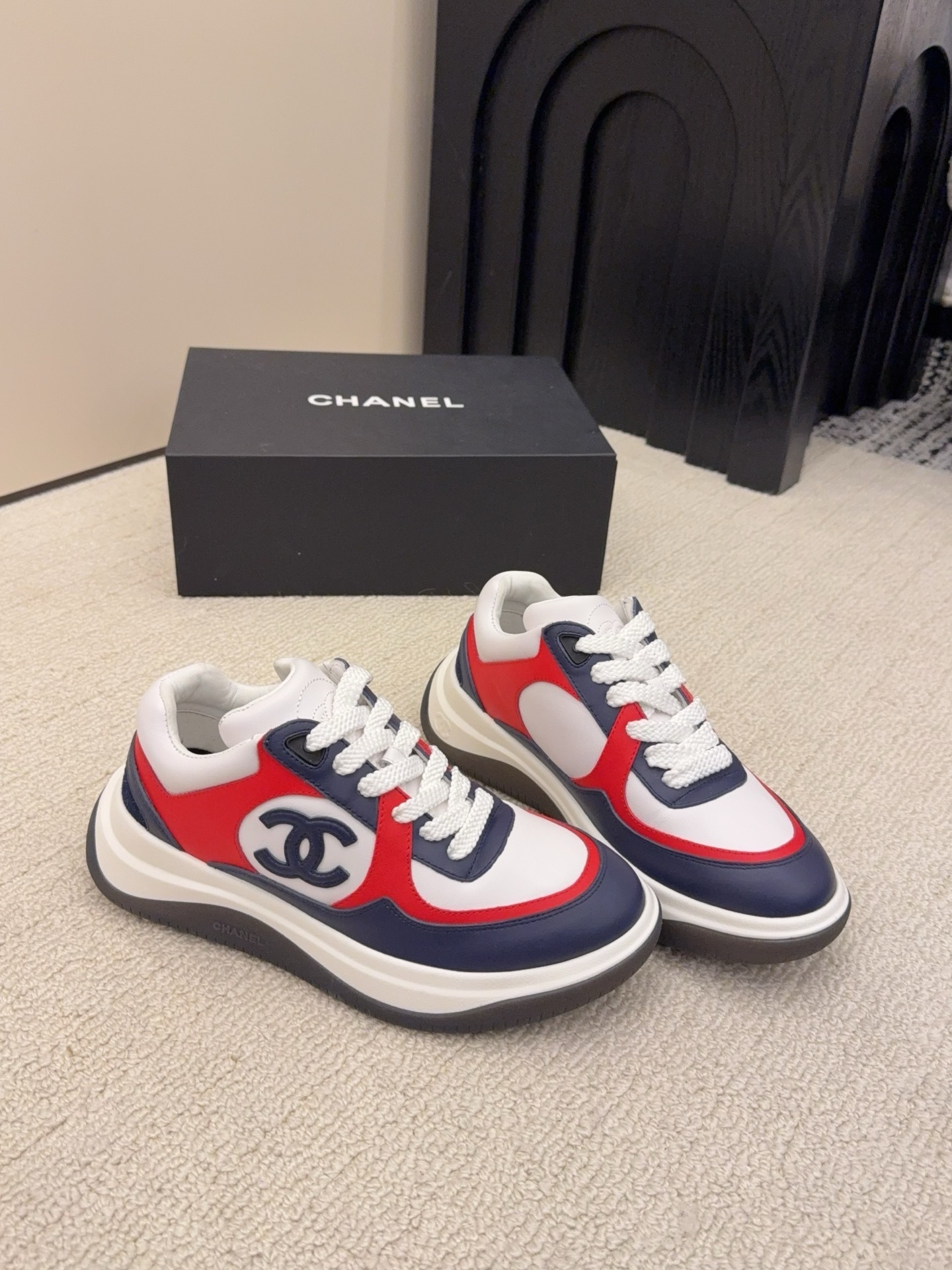 A pair of Chanel tricolor sneakers in white, navy blue, and red with a chunky sole and the CC logo, displayed next to a black Chanel shoe box. - Hình 8